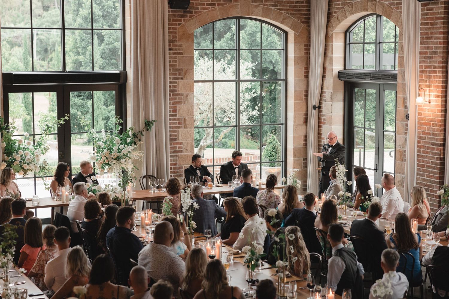 Wedding guests dine at long tables inside a rustic brick venue with tall arched windows and natural lighting.