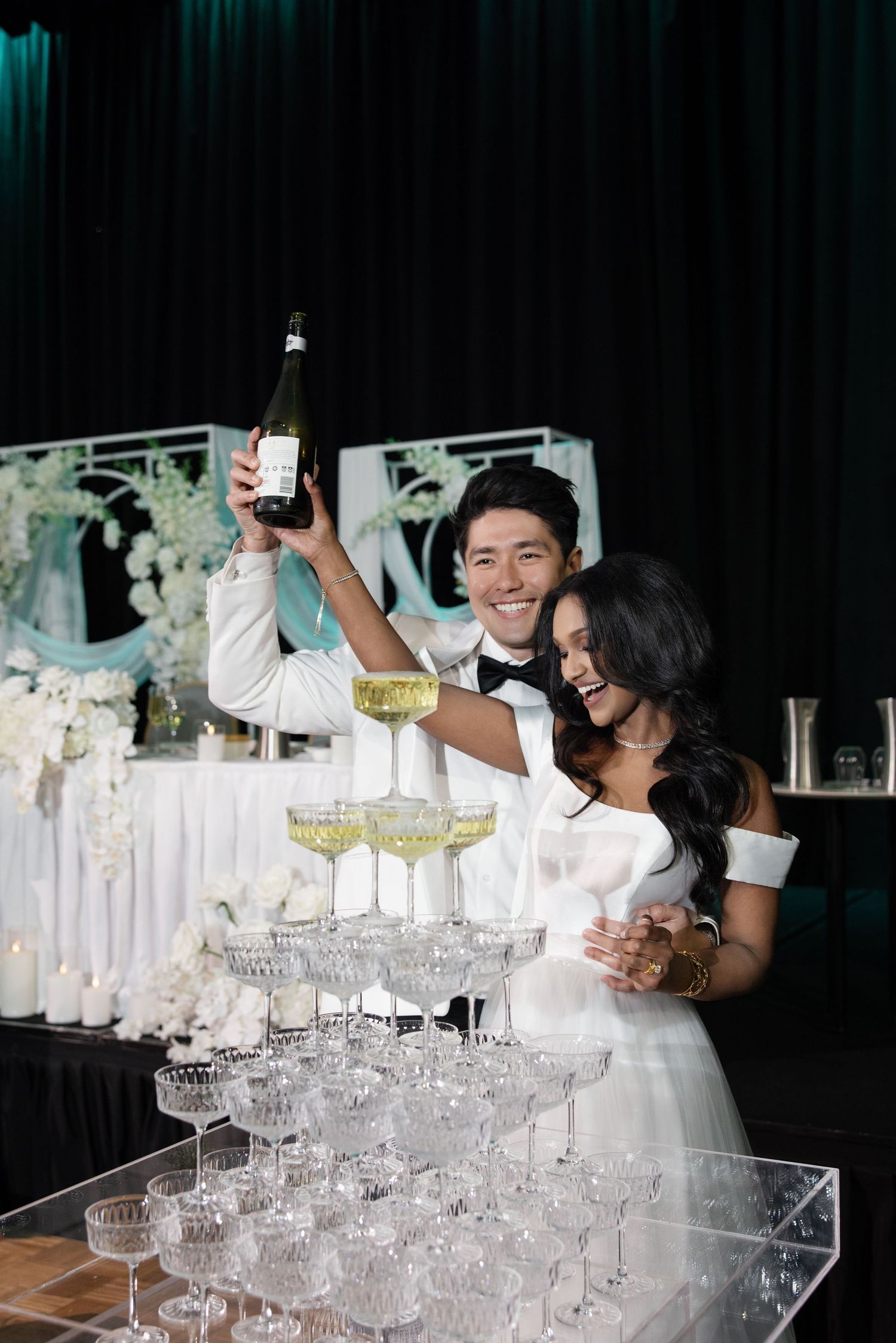 A champagne tower being poured at an elegant wedding reception with crystal glasses stacked in multiple tiers.