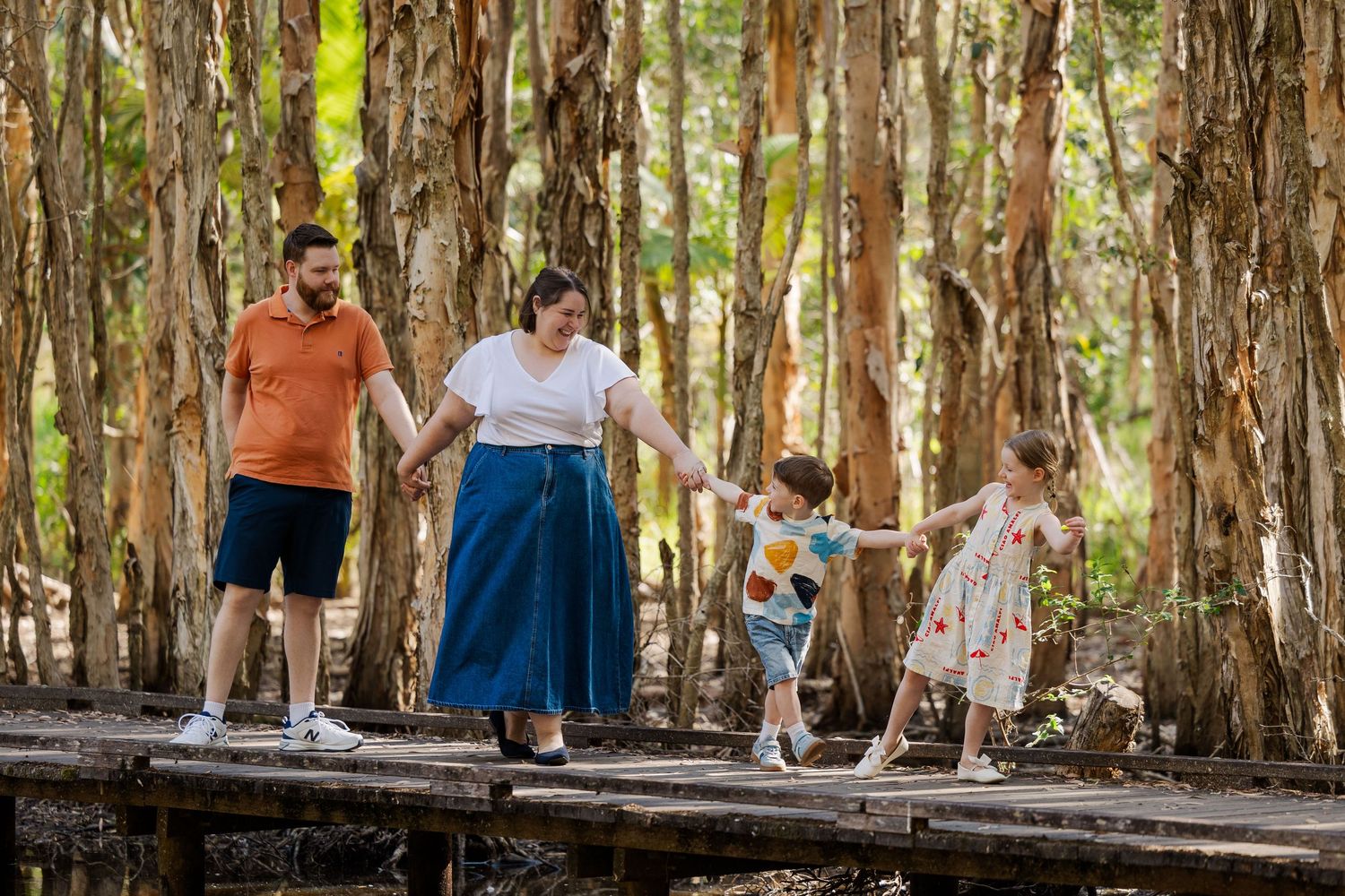 A family walks together holding hands on a wooden boardwalk path through a eucalyptus forest.