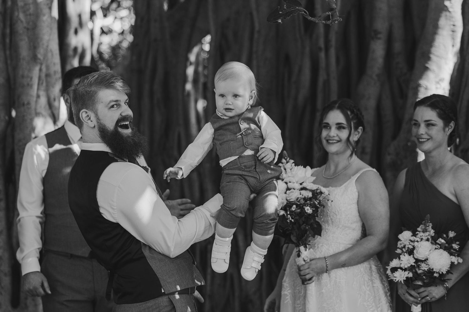 Black and white wedding photo of bearded groom lifting baby while bride holds bouquet with bridesmaid outdoors.