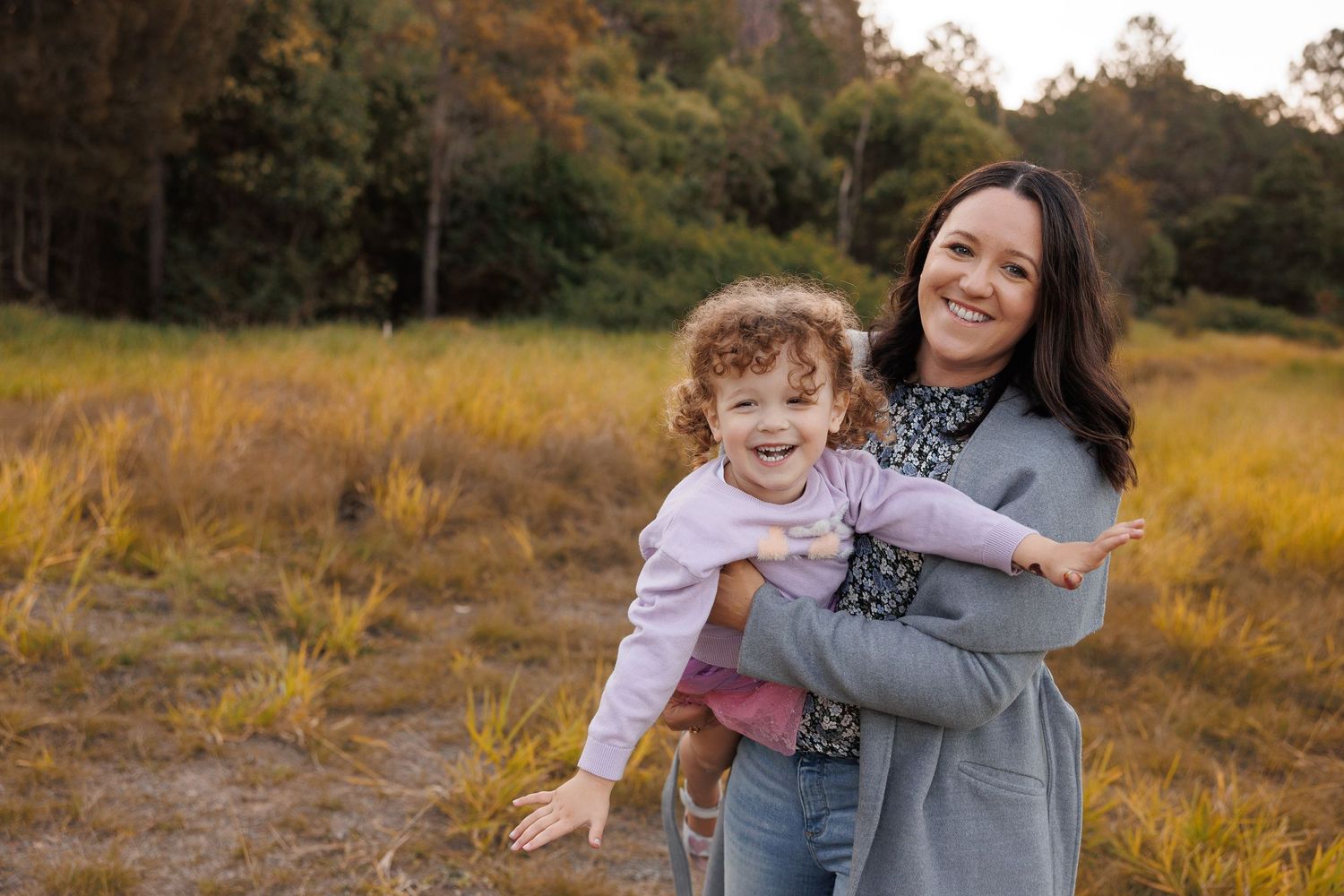 A smiling mother holds her curly-haired toddler in a sunny autumn field with golden grass and trees.
