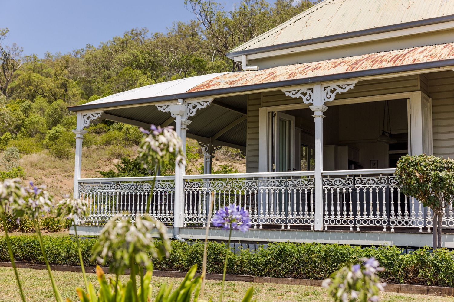 Charming white Victorian cottage with ornate verandah railings, surrounded by lush gardens and rolling hills.