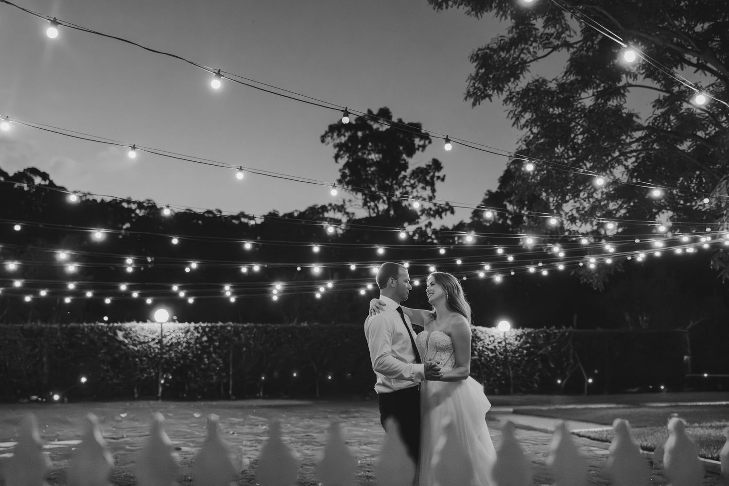 Black and white photo of a couple sharing a romantic kiss under string lights at an outdoor evening wedding reception.