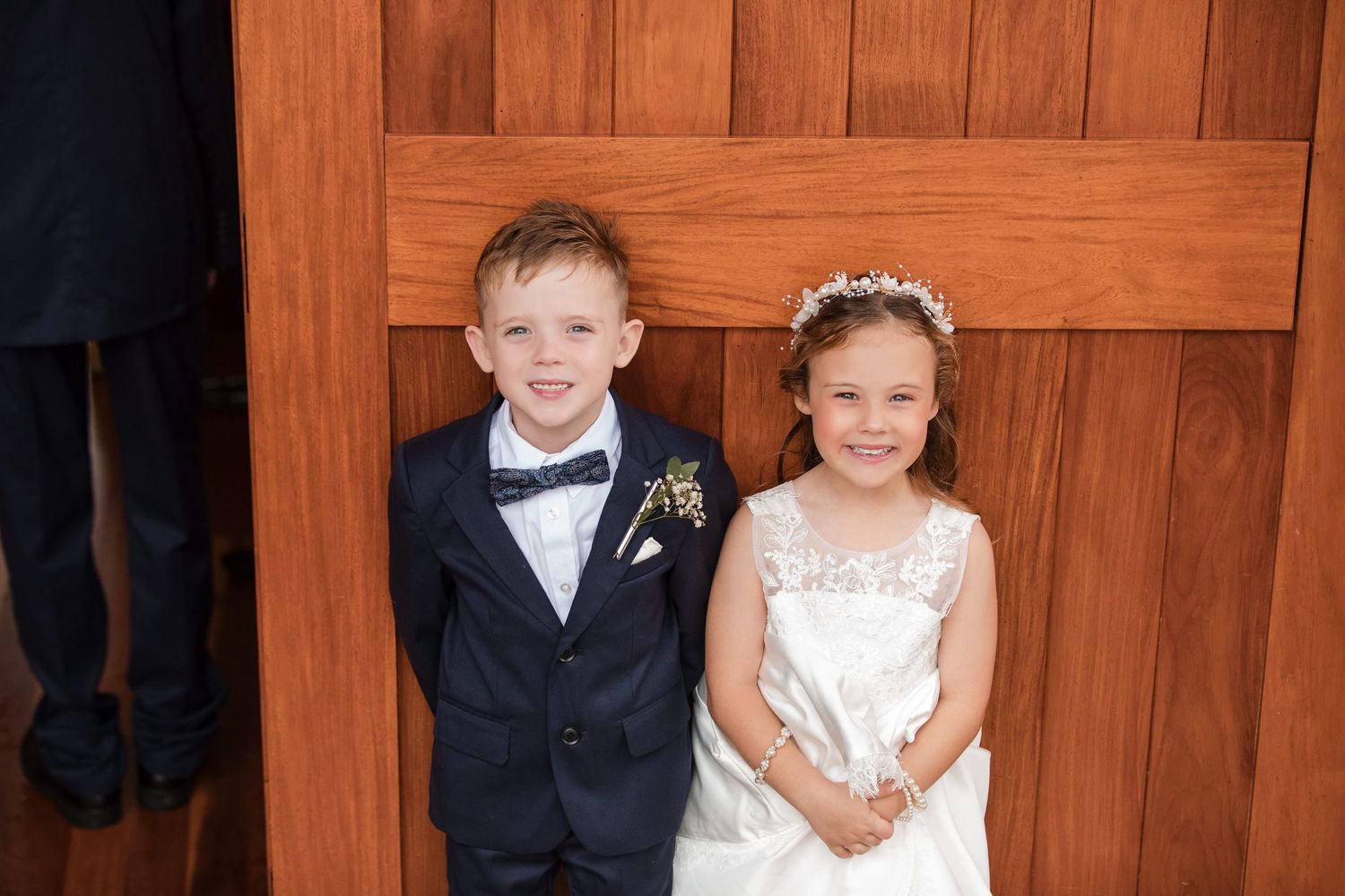 Young ring bearer in navy suit and flower girl in white dress smiling at a wedding ceremony.