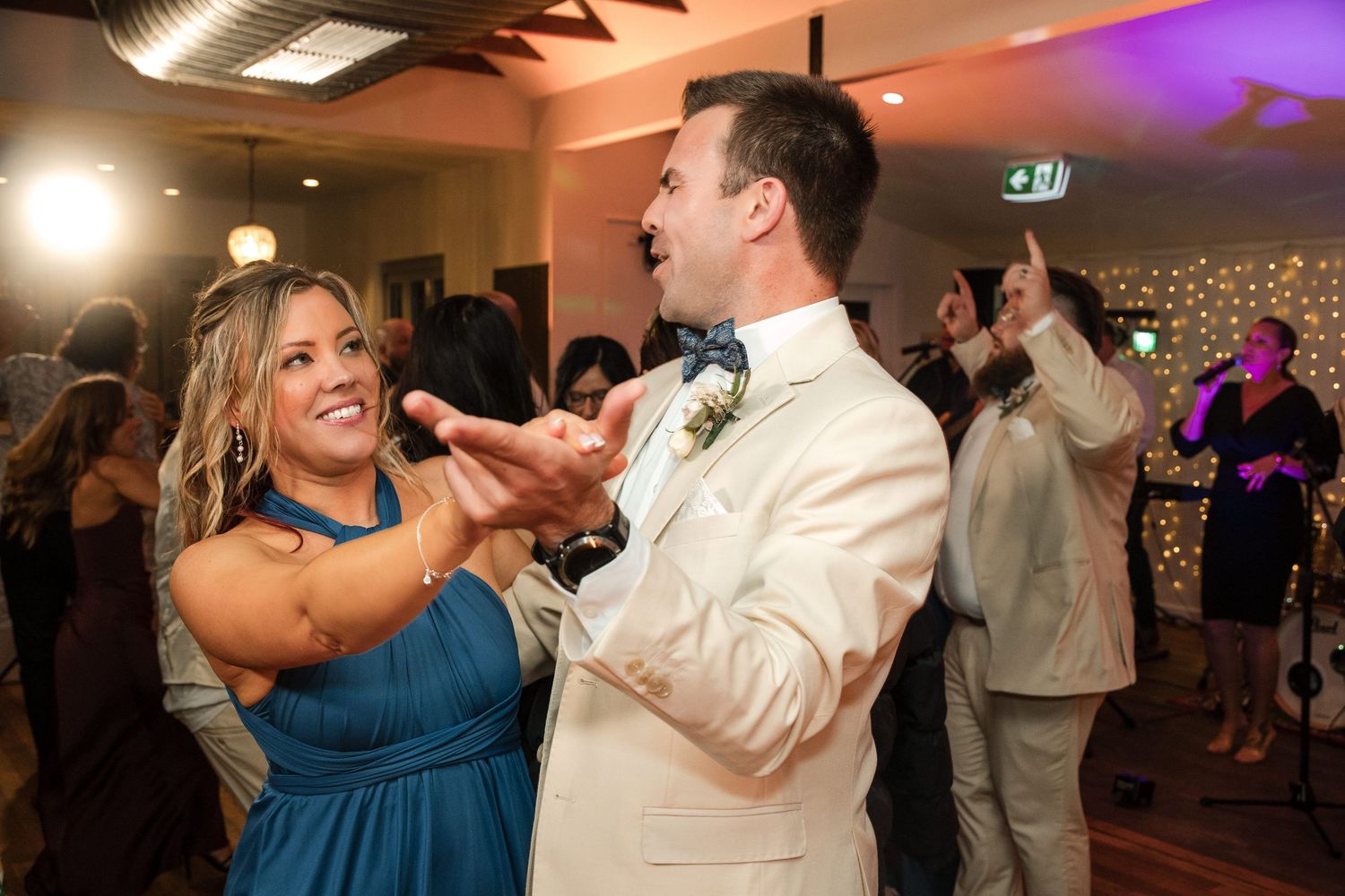 Bride and groom dancing joyfully at wedding reception, woman in teal dress, man in white suit with boutonniere.