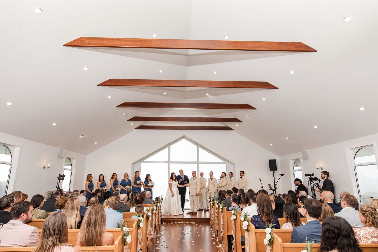 Elegant wedding ceremony in a white chapel with wooden beam ceiling, bridal party at altar, guests seated in pews.