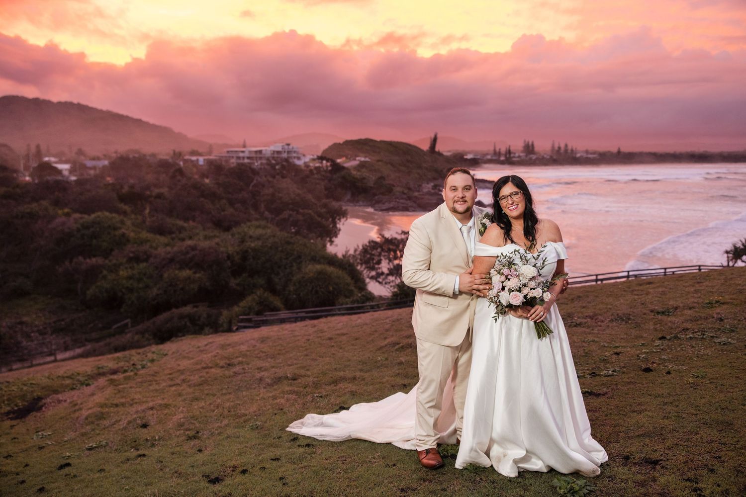 Bride and groom pose at sunset overlooking coastal scenery with pink sky and ocean views at their beach wedding.