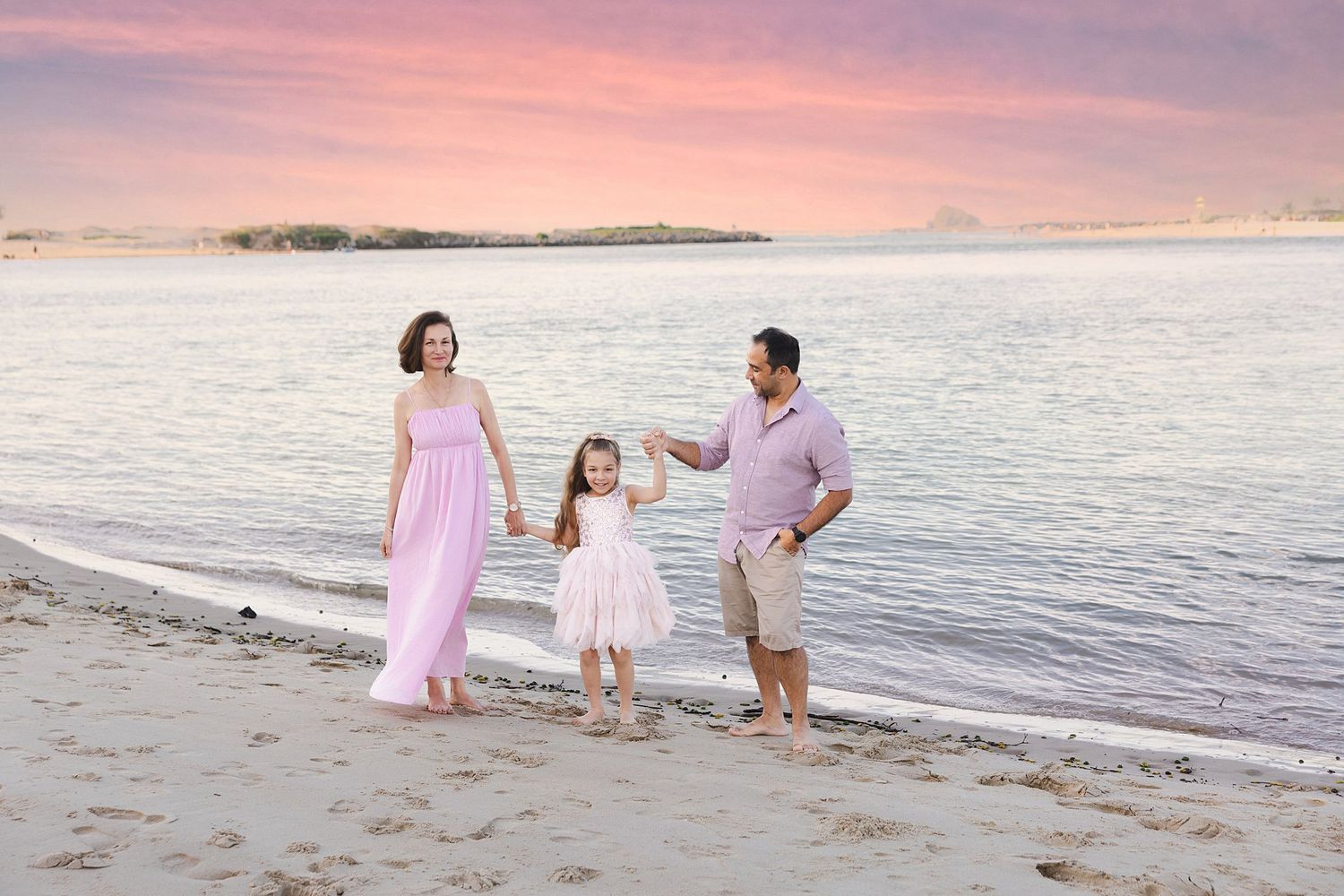 Three people holding hands and walking along a beach at sunset with pink sky and calm water in the background.