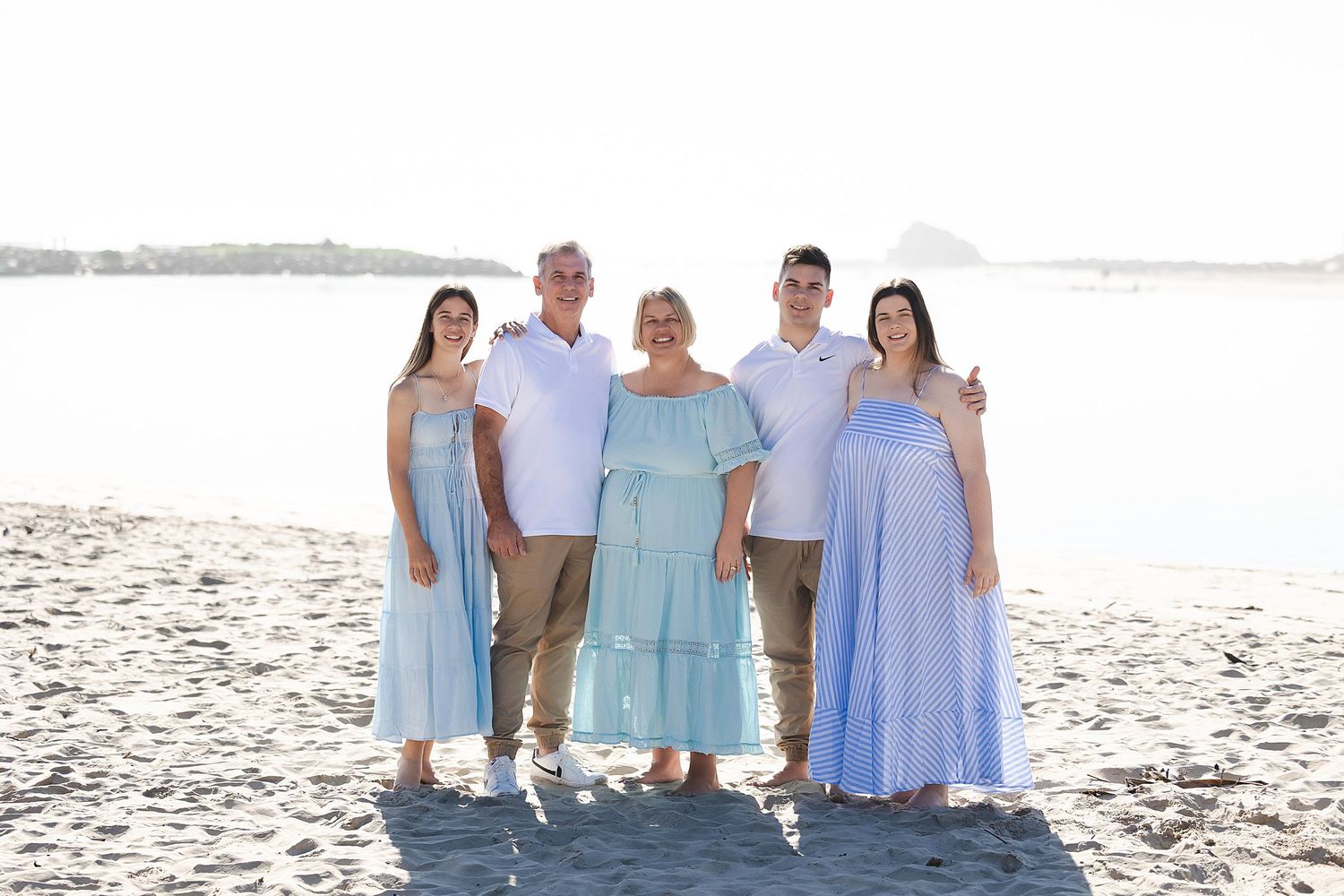 A family in coordinating light blue outfits poses together on a sunny beach with water in the background.