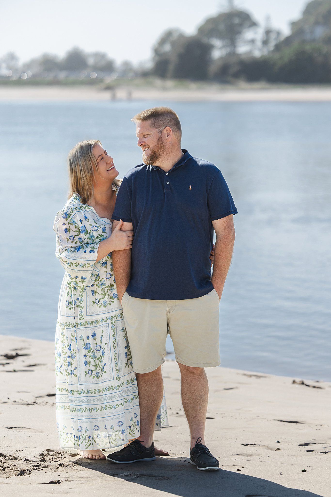 A couple shares a tender moment while standing on a beach with calm water and trees in the background.