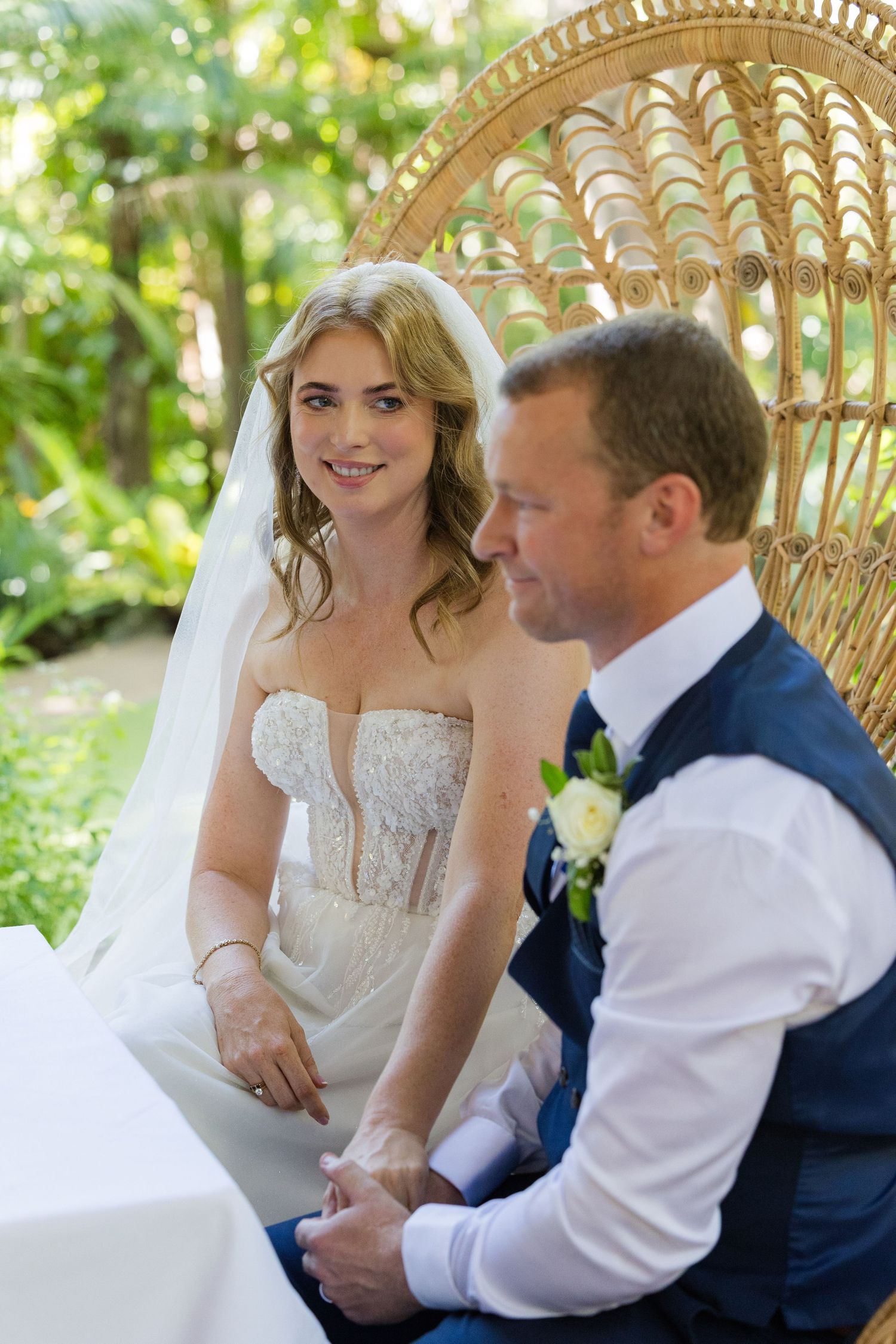 Romantic wedding couple shares intimate moments while seated in a decorative wicker peacock chair in a garden setting.