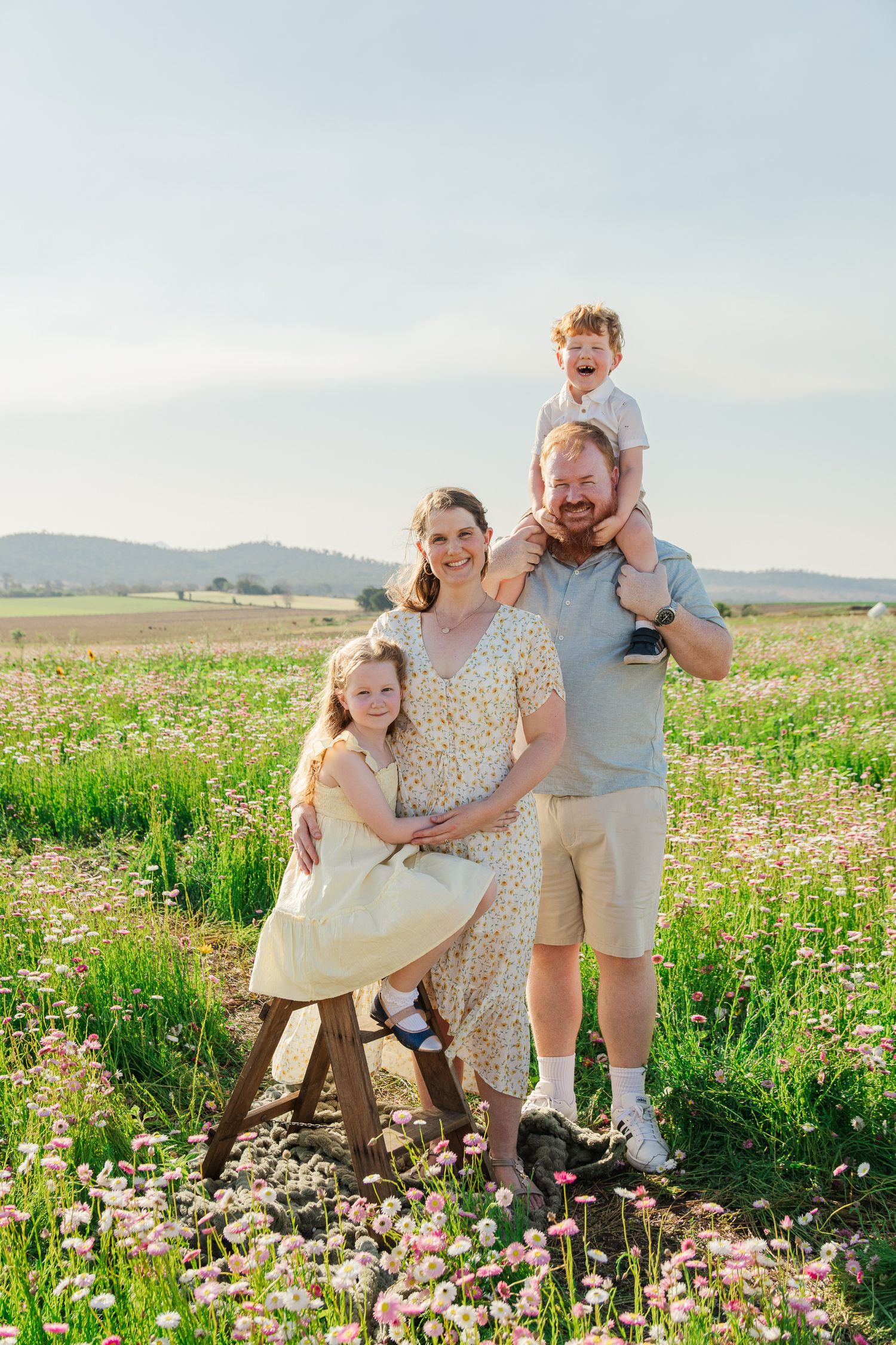 Happy family of four posing in a blooming wildflower meadow on a sunny day, with hills in the background.