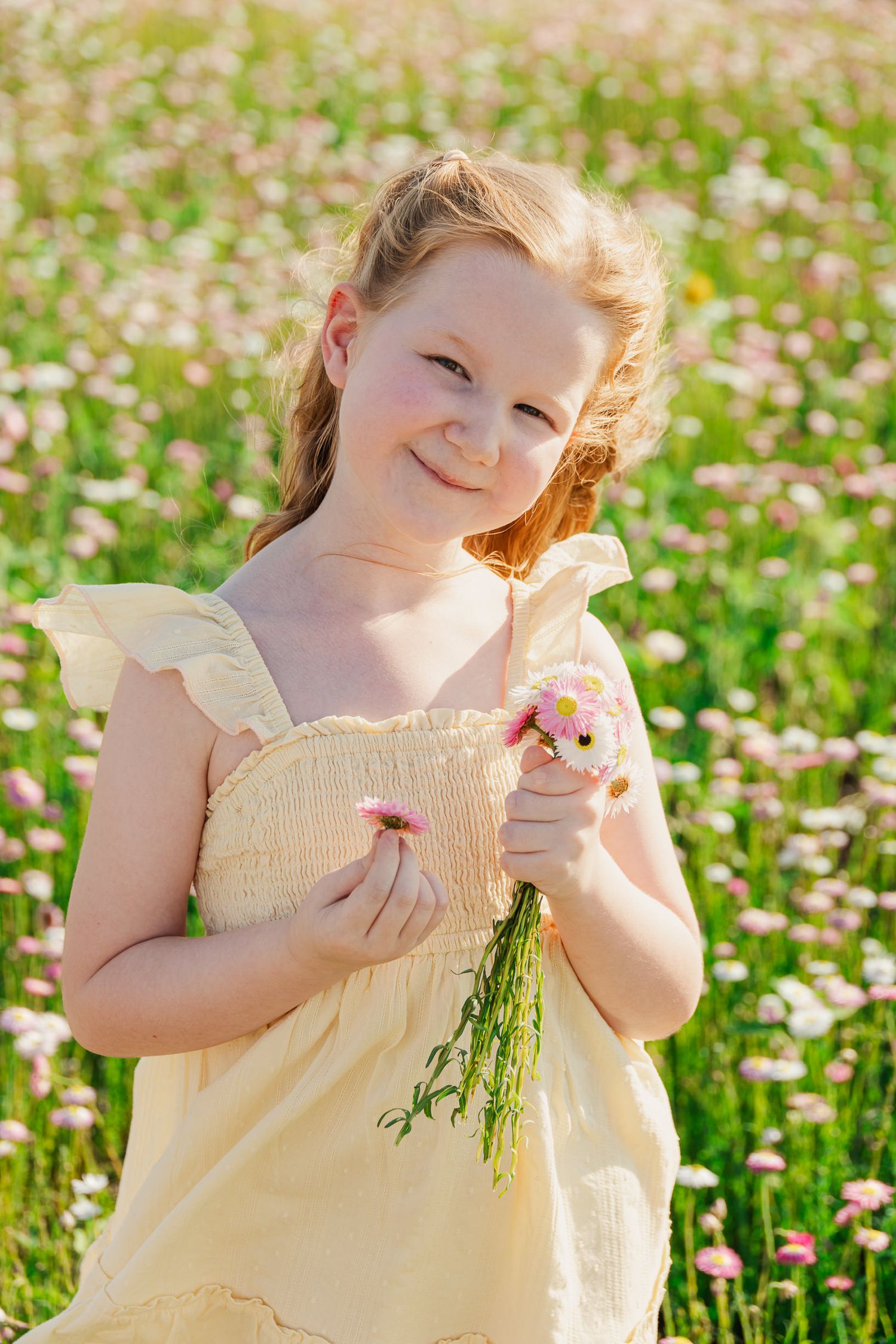 Smiling young girl in yellow dress holding pink wildflowers in a sunny blooming meadow field.