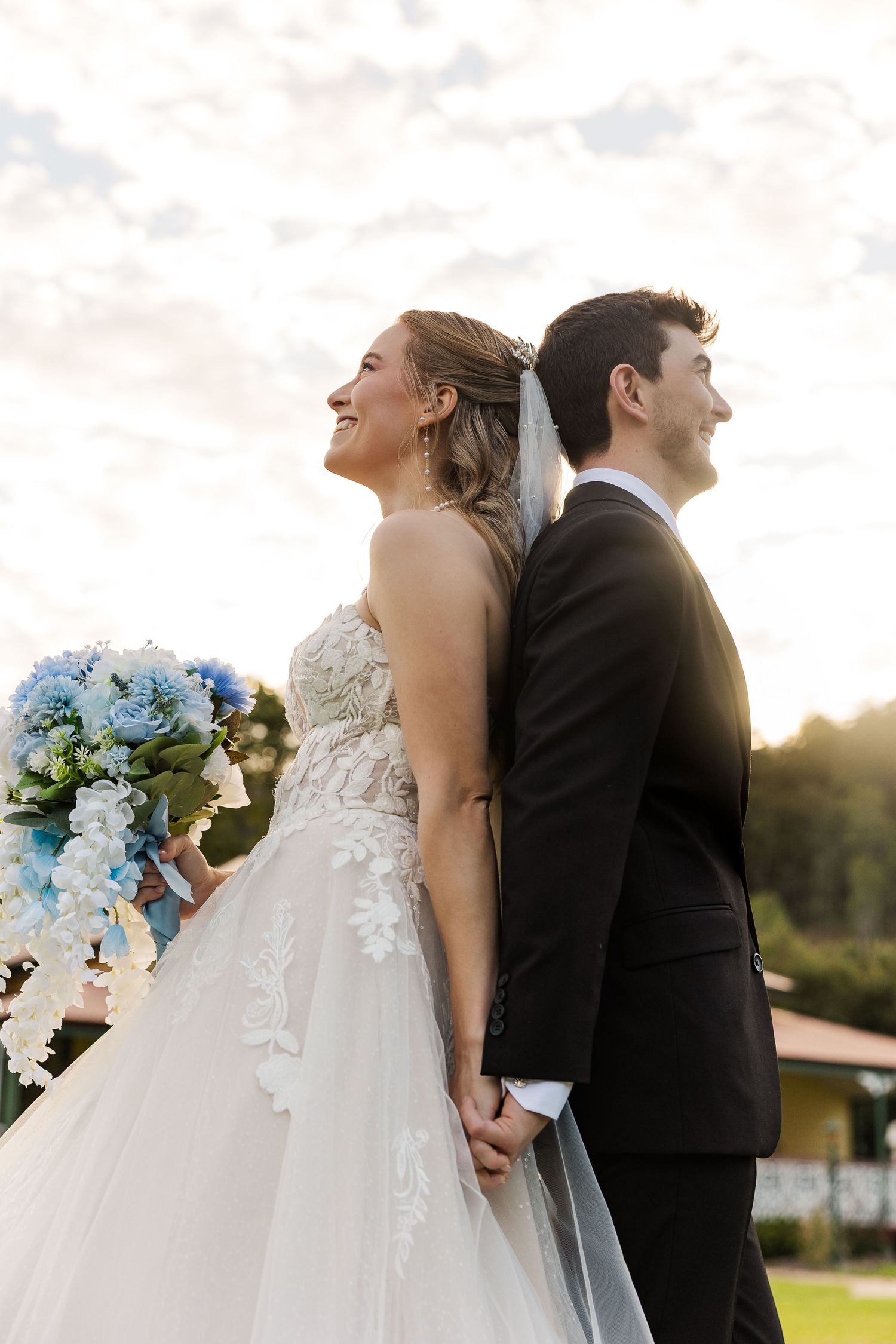 Bride and groom stand back-to-back outdoors, she holds blue bouquet, both smiling, dressed in wedding attire at sunset.