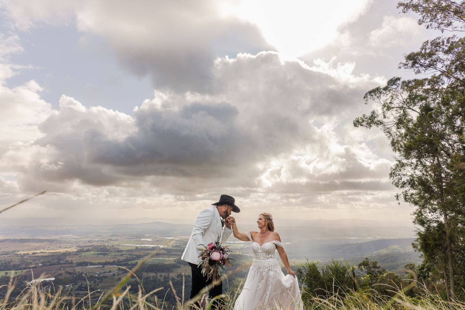Bride and groom share a romantic moment on a scenic hilltop with dramatic clouds and sweeping valley views behind them.