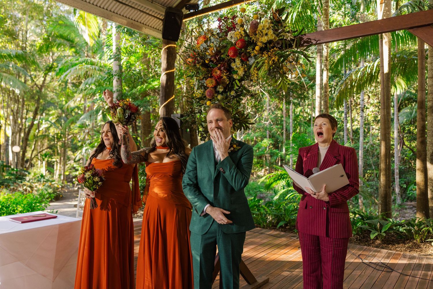 Groom in green suit laughs with bridesmaids in rust dresses and celebrant in maroon at outdoor wedding.