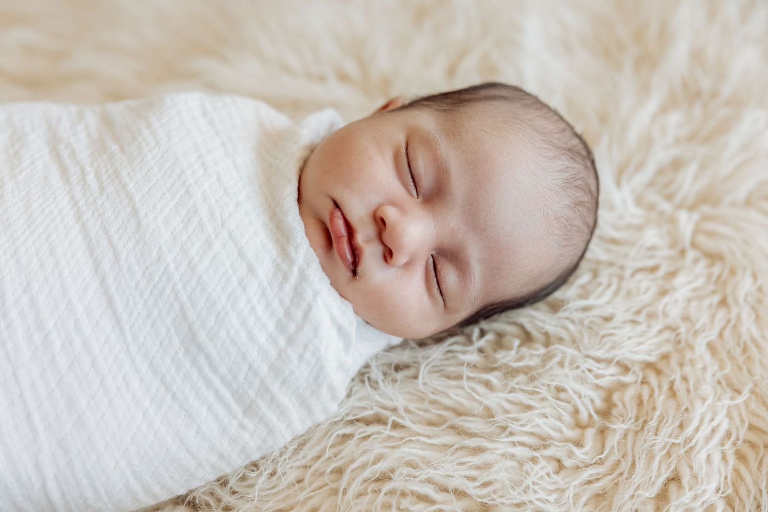 Peaceful sleeping newborn baby swaddled in white blanket resting on soft cream textured fabric.