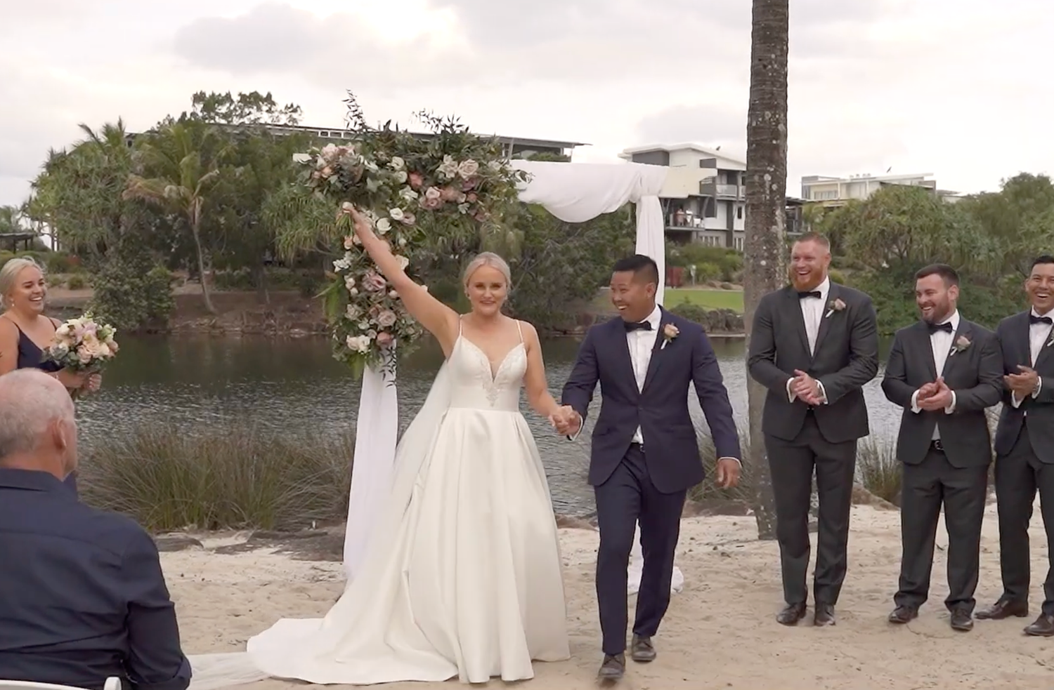 A wedding party walks along a waterfront path as the bride raises her bouquet in celebration during an outdoor ceremony.
