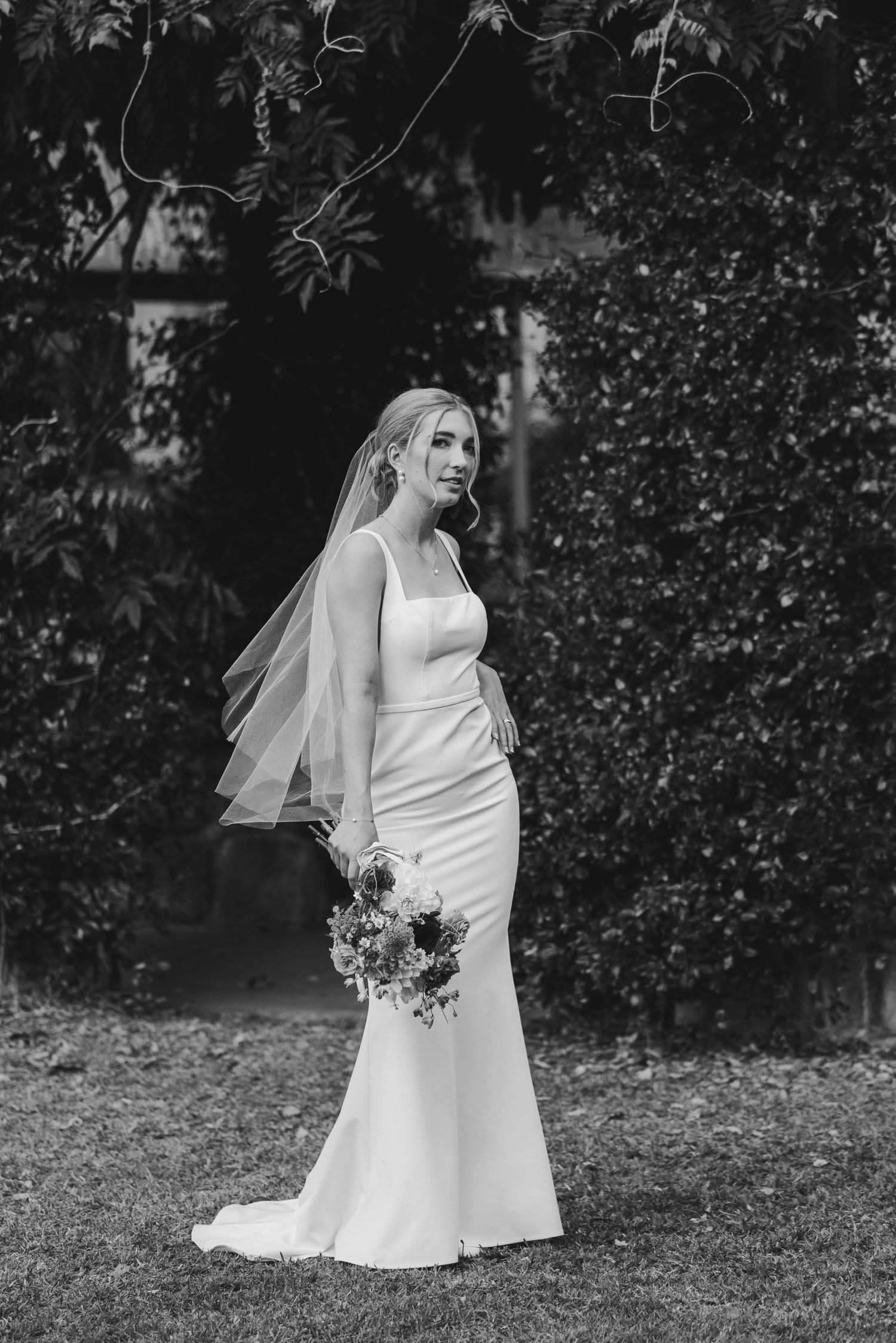 A bride in a classic white wedding dress and veil poses elegantly in a garden setting with a floral bouquet.