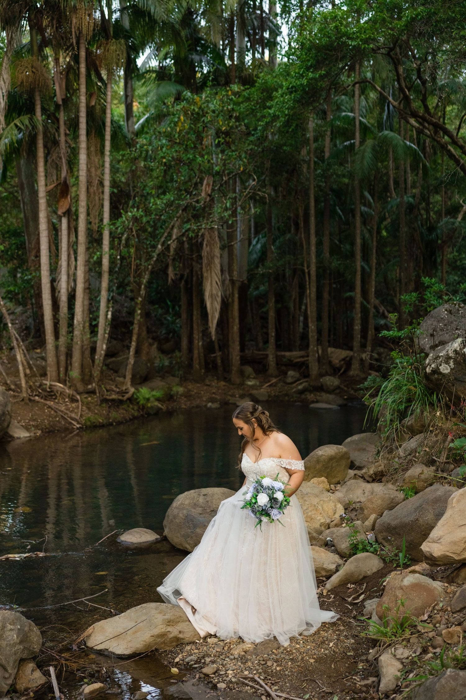 A person in a white wedding dress stands beside a tranquil forest pond surrounded by tall trees and rocks.