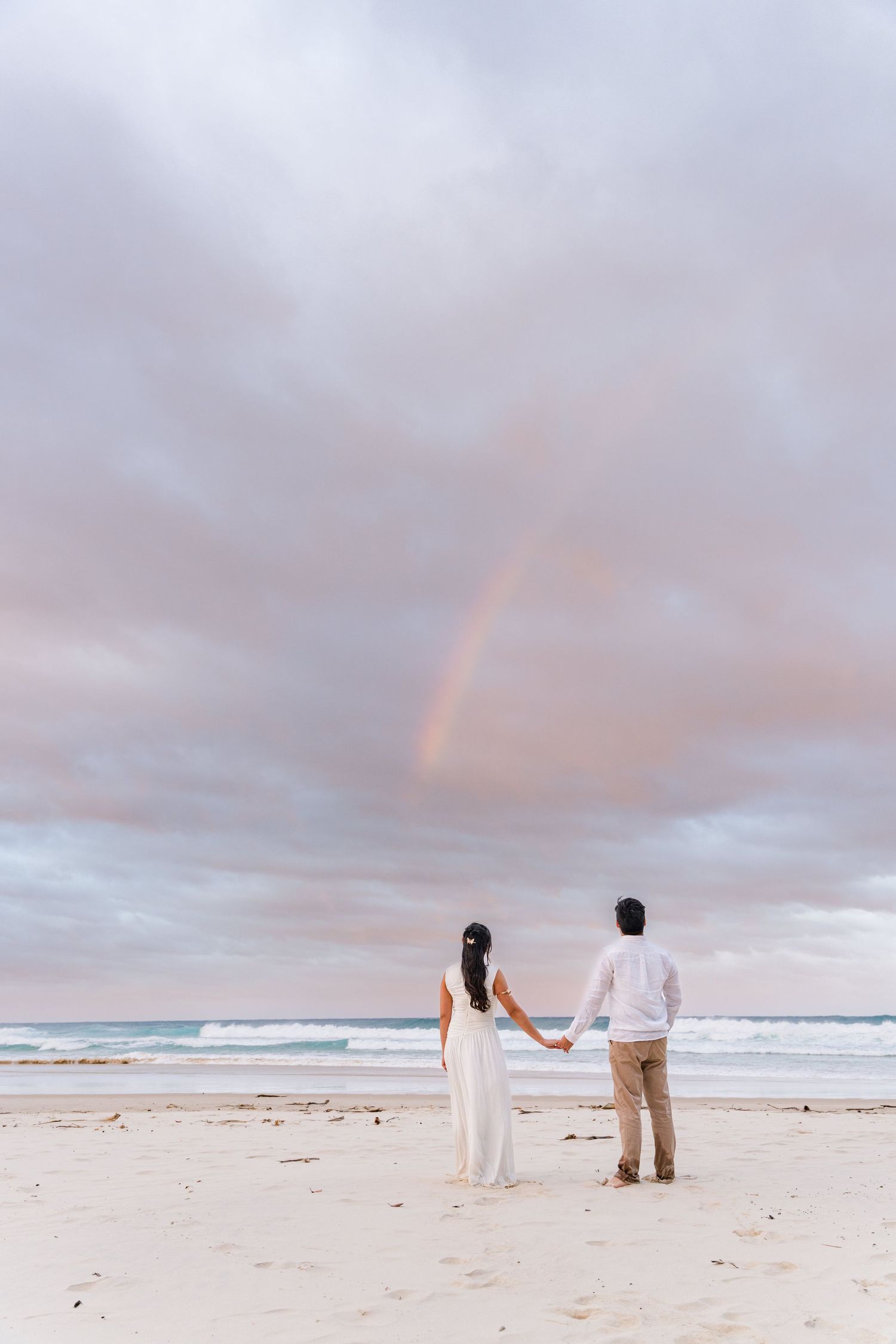 Couple holding hands on a beach in wedding attire, gazing at a rainbow over the ocean under a cloudy sky.