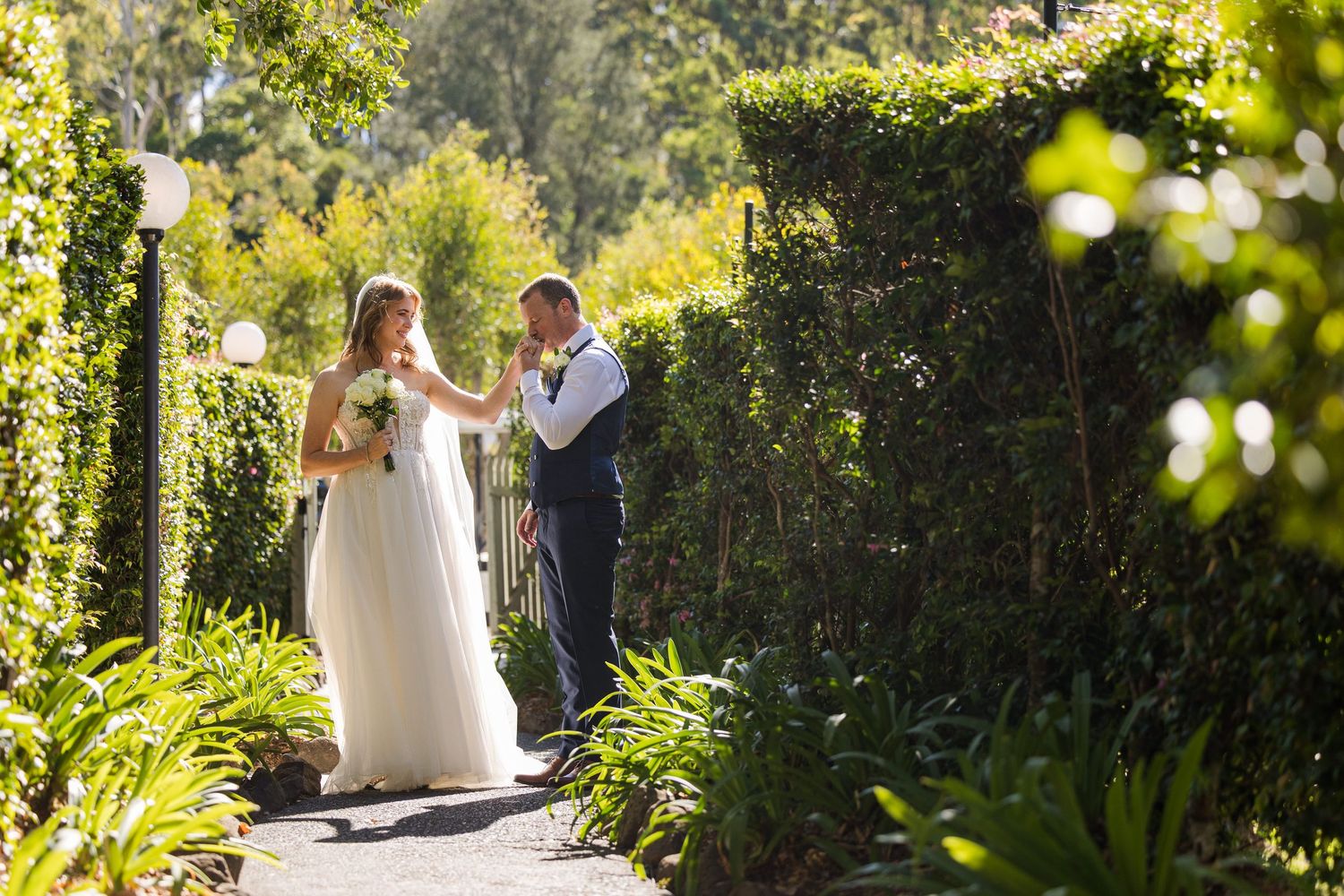 Newlywed couple walking along lush garden path surrounded by greenery in romantic outdoor setting.