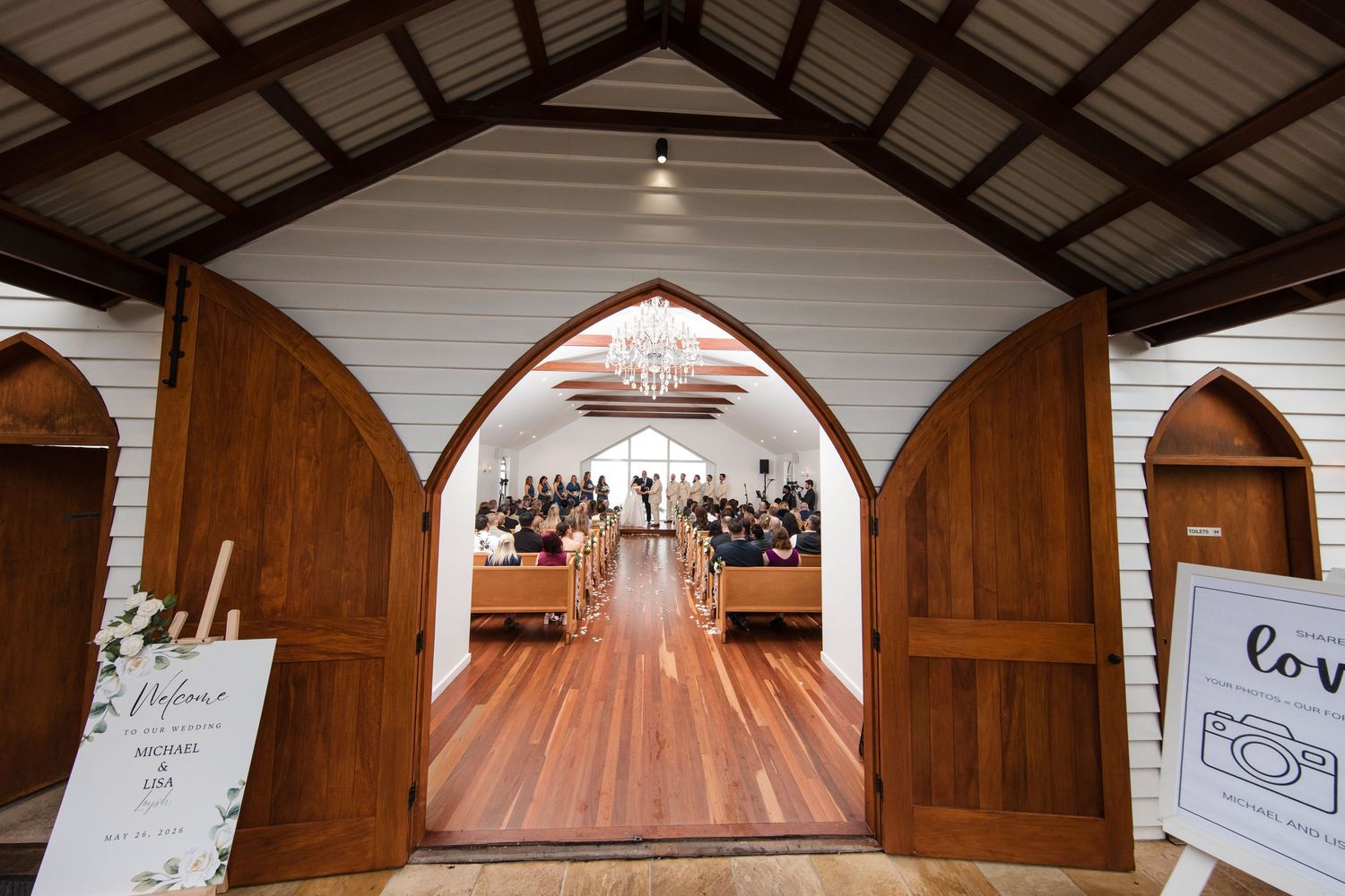 Rustic white chapel wedding venue with open wooden doors revealing guests seated along a polished timber aisle.