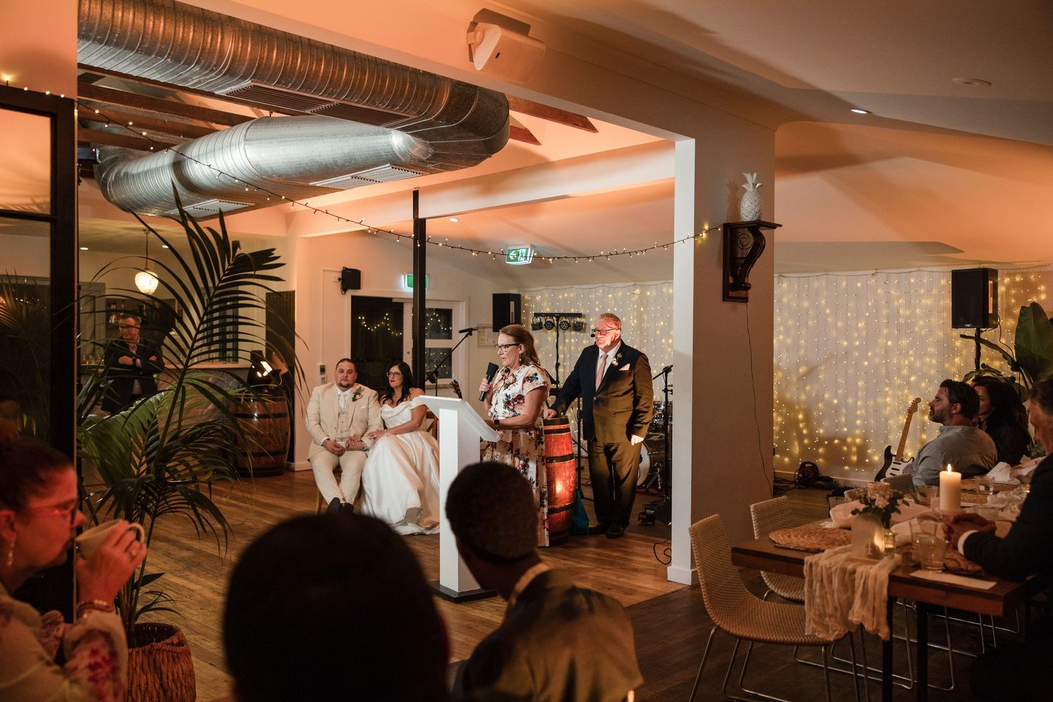 Wedding reception with couple seated on stage, live band performing, guests watching in warmly lit venue.