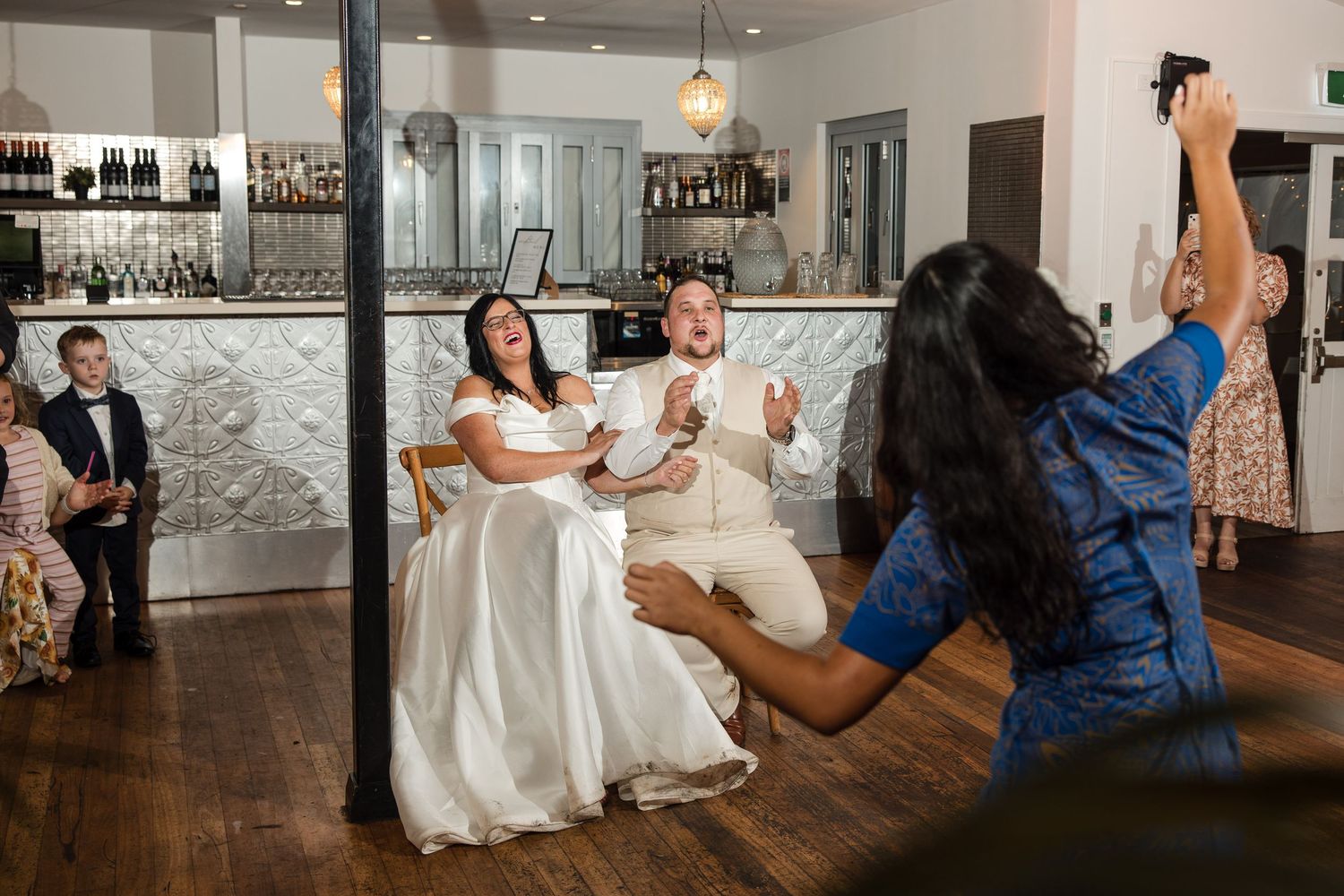 Bride and groom seated during wedding reception game while guest photographs them at elegant bar venue.