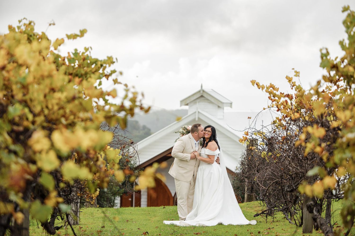 Bride and groom embracing in a lush vineyard with a white chapel and mountains in the background.