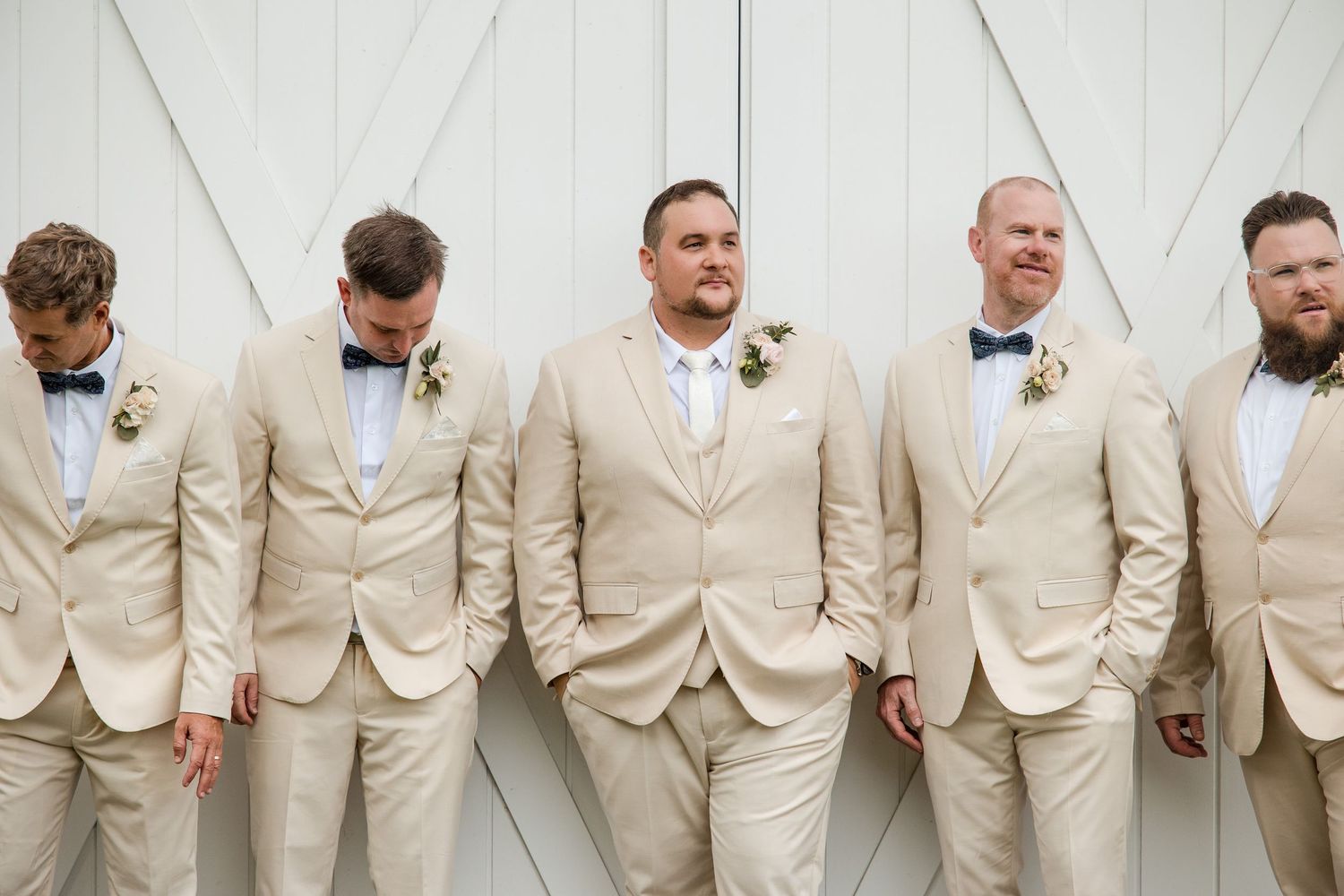 Five groomsmen in matching tan suits with navy bow ties and boutonnieres pose casually against white barn doors.