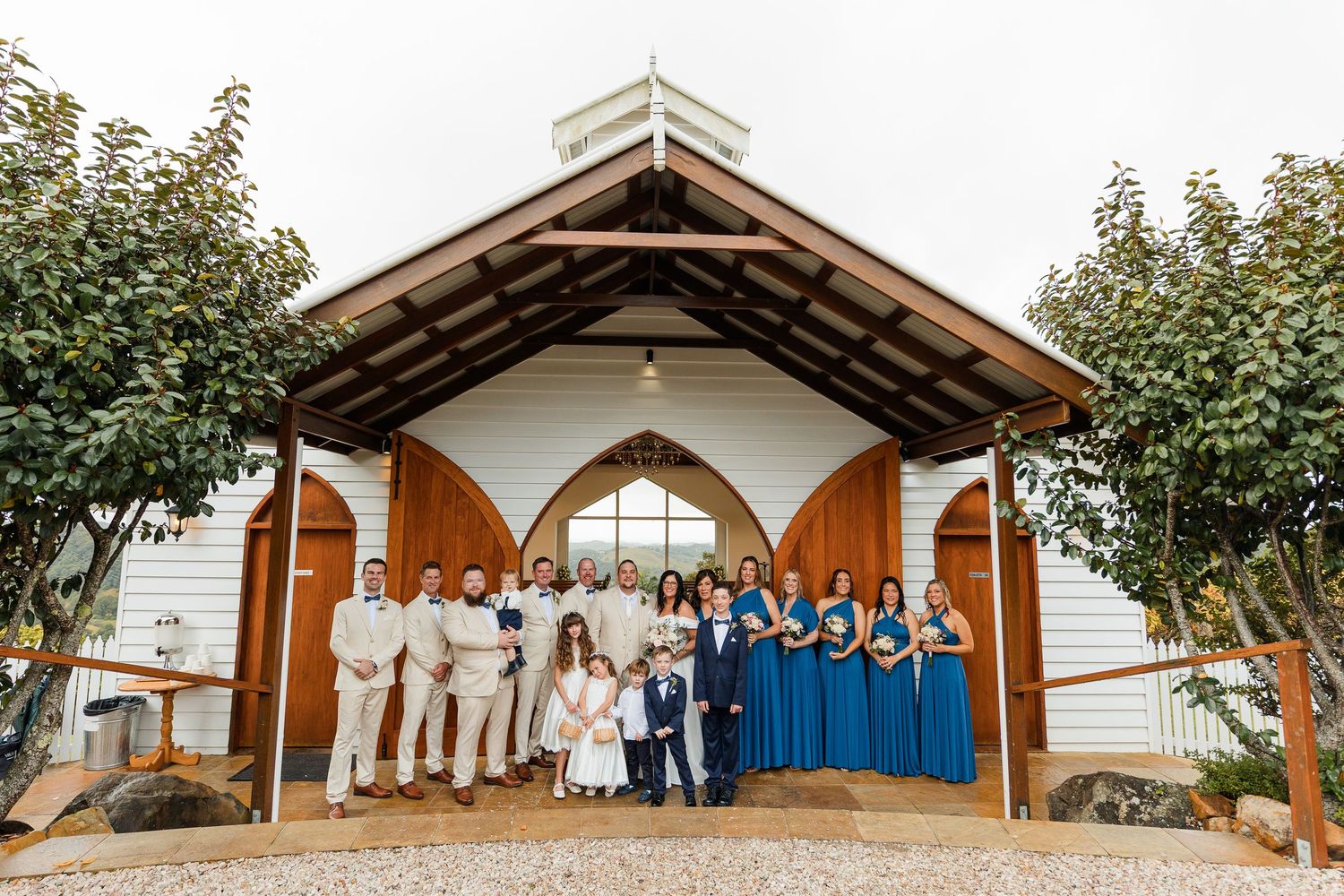 Wedding party poses before a white chapel with wooden doors, groomsmen in tan suits and bridesmaids in blue dresses.