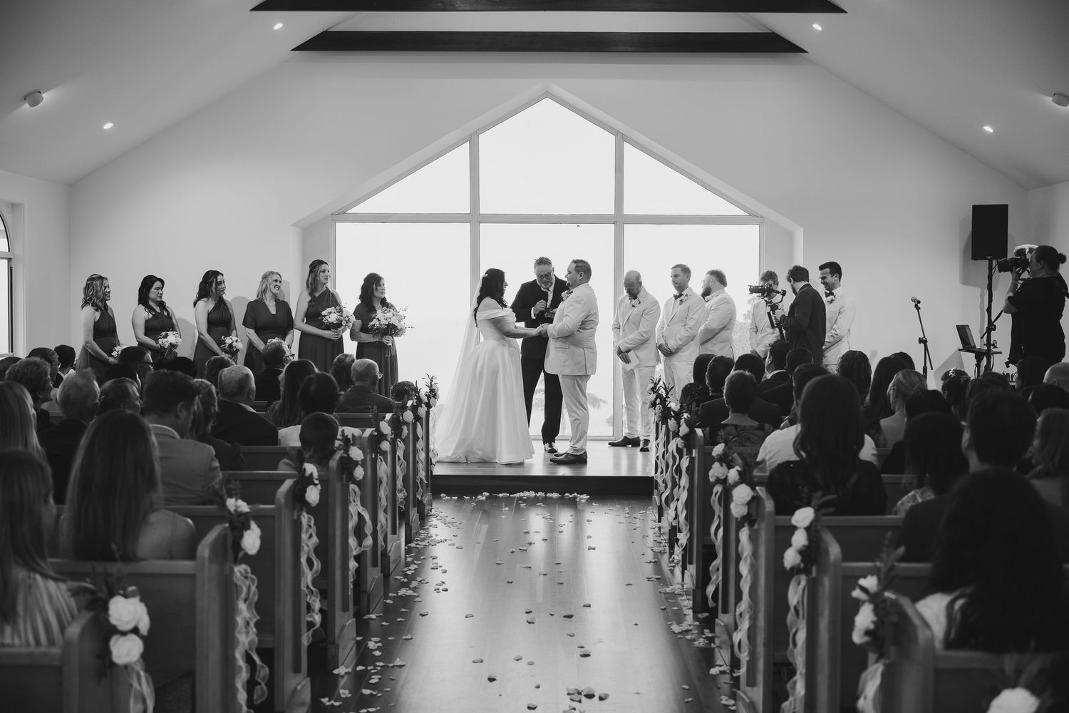 Black and white wedding ceremony photo inside a chapel with bride and groom at altar surrounded by wedding party.