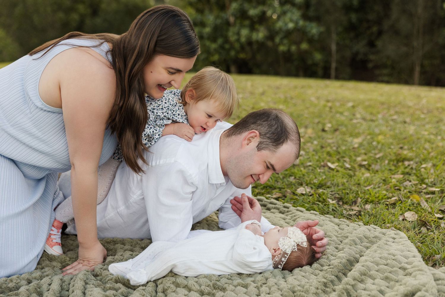 Family of four admiring newborn baby on blanket outdoors, parents and toddler bonding in park setting.