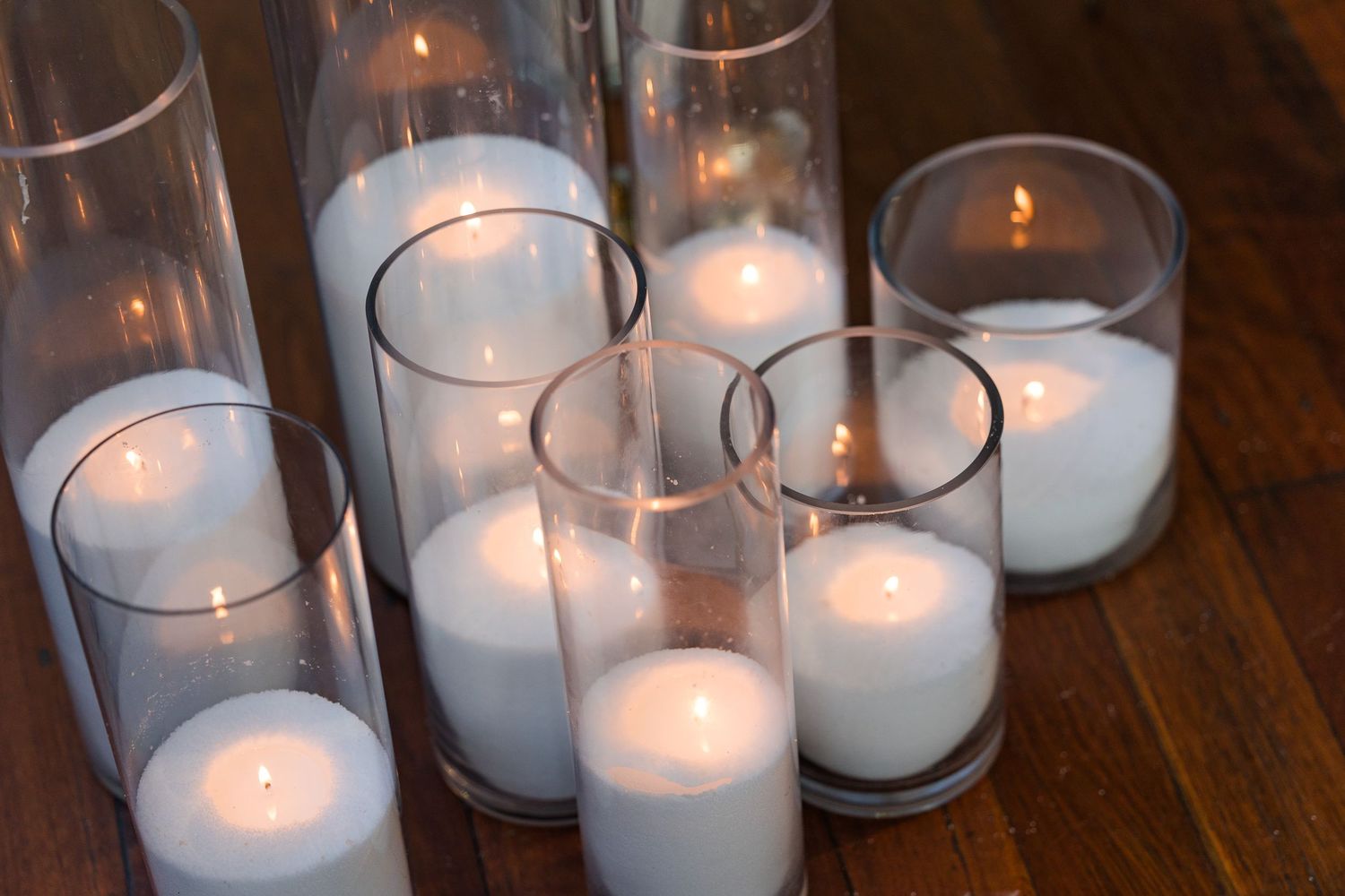 Multiple white pillar candles glowing in tall and short clear glass hurricane vases arranged on a wooden floor.
