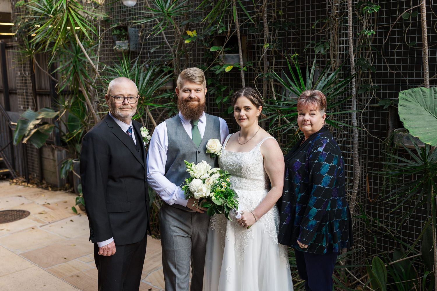 Bride and groom pose with family members in an indoor garden setting, bride holding white bouquet.
