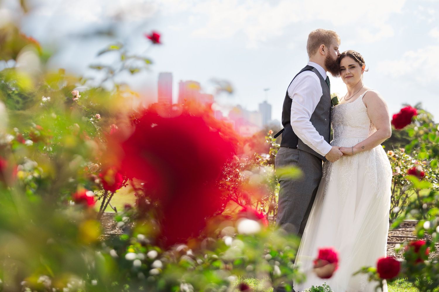 Bride and groom embracing in a lush rose garden with red blooms and city skyline in the background.