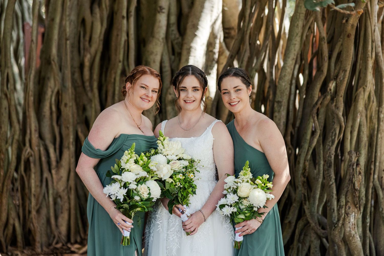 Bride in white lace gown poses with two bridesmaids in sage green dresses holding white bouquets before large banyan tree.