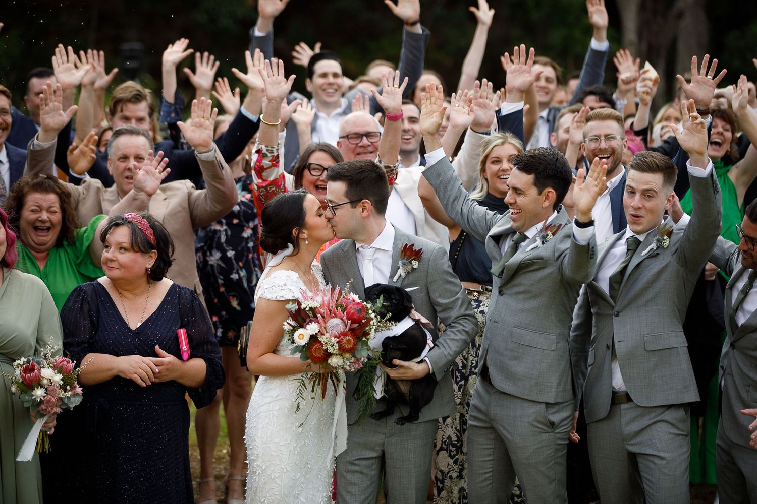 Joyful wedding party celebrates with hands raised as bride and groom kiss, holding a dog, outdoors.