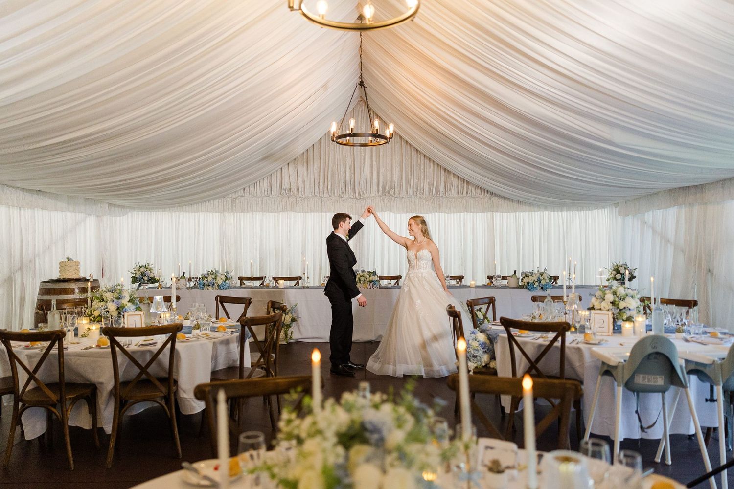 Bride and groom share first dance in elegant white draped wedding tent with floral centerpieces and candlelit tables.