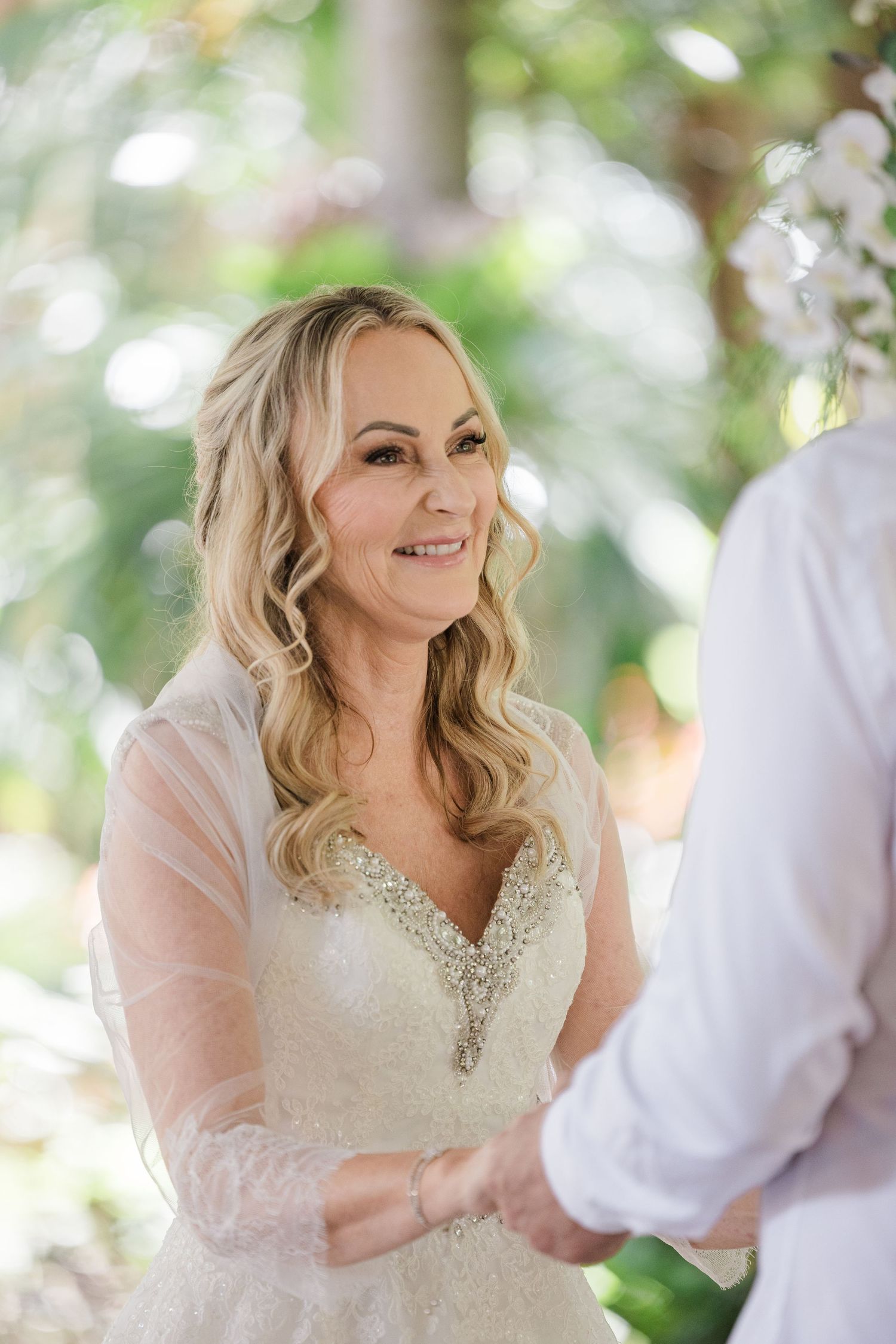 Bride in a beaded sweetheart gown smiles warmly at groom during outdoor wedding ceremony.