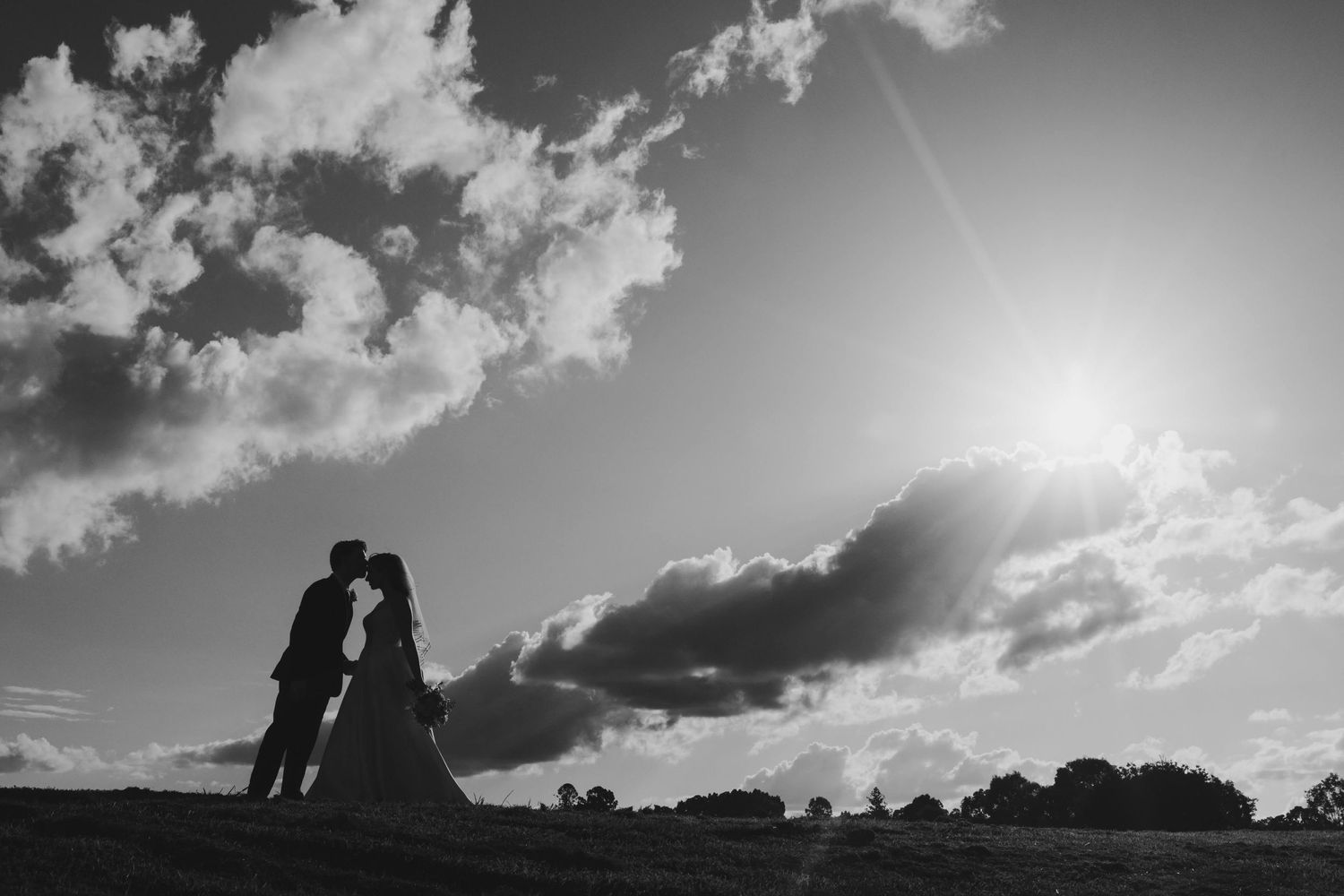 Silhouettes of a couple embracing against a dramatic sunset sky with clouds and sunburst.