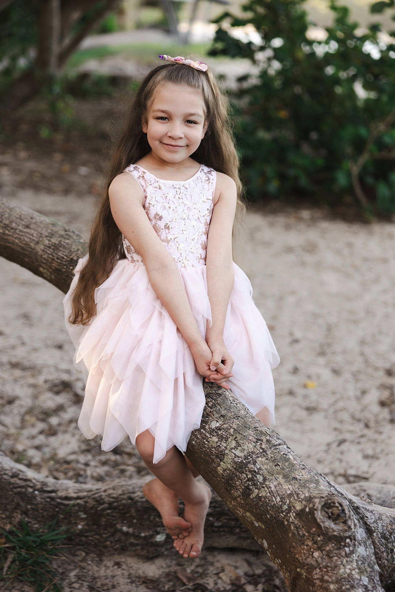 Child in pink tulle dress with lace bodice sits barefoot on fallen tree trunk outdoors.