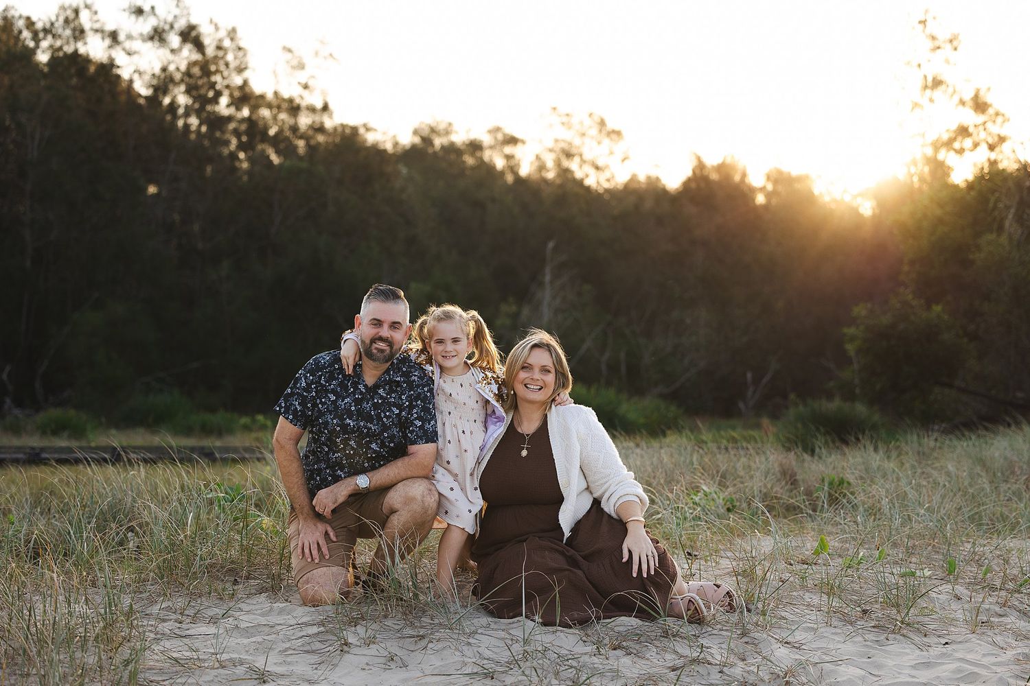 A family sits together on dry grass during sunset with trees in the background.