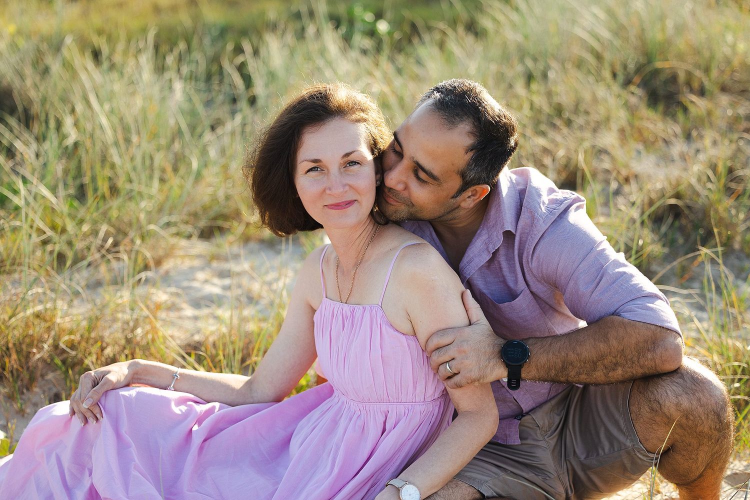 A couple in coordinating purple outfits share an intimate moment together in a sunlit meadow.