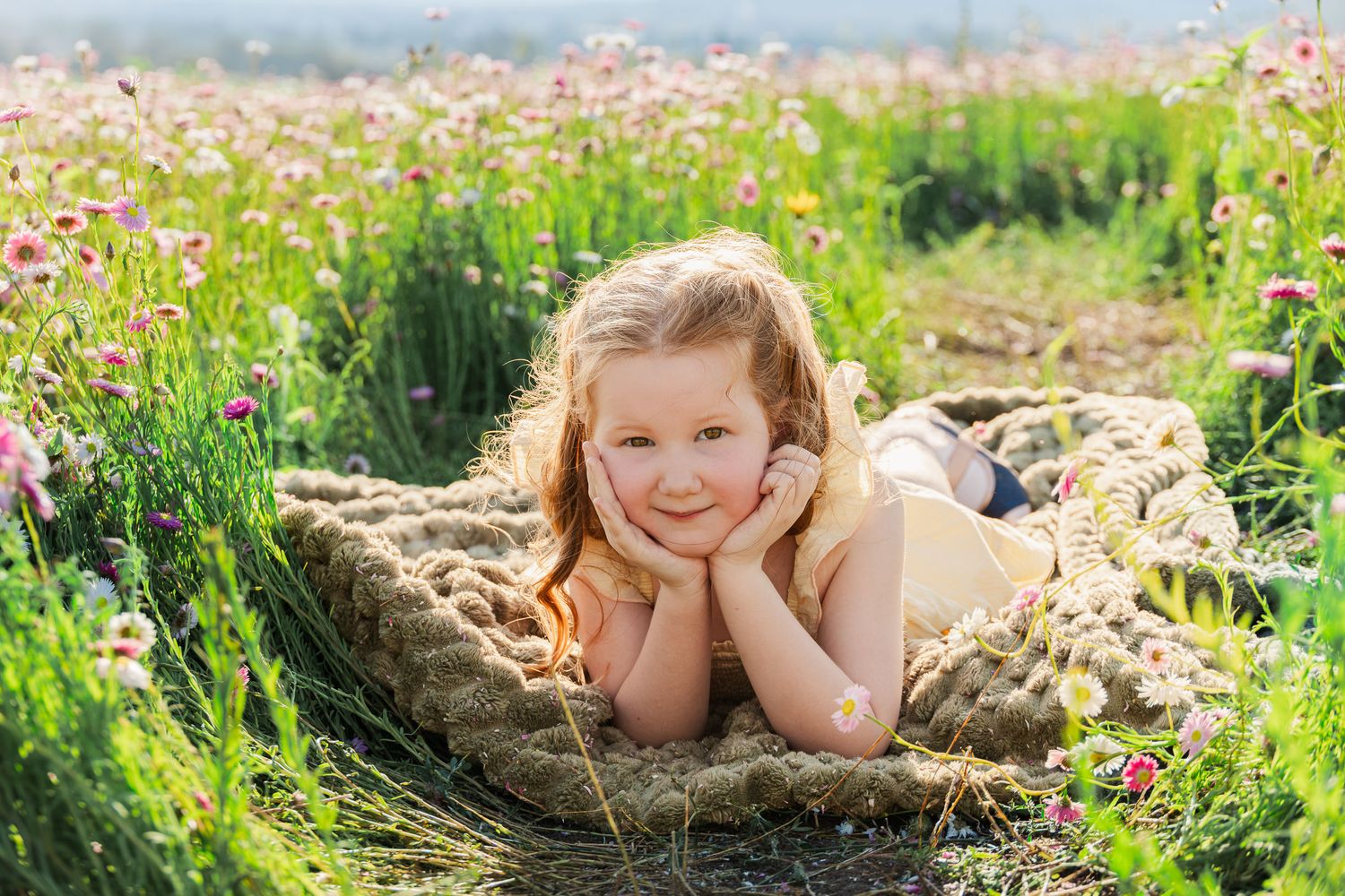 A young girl with blonde hair lies on a blanket in a blooming flower field, smiling with hands on cheeks.