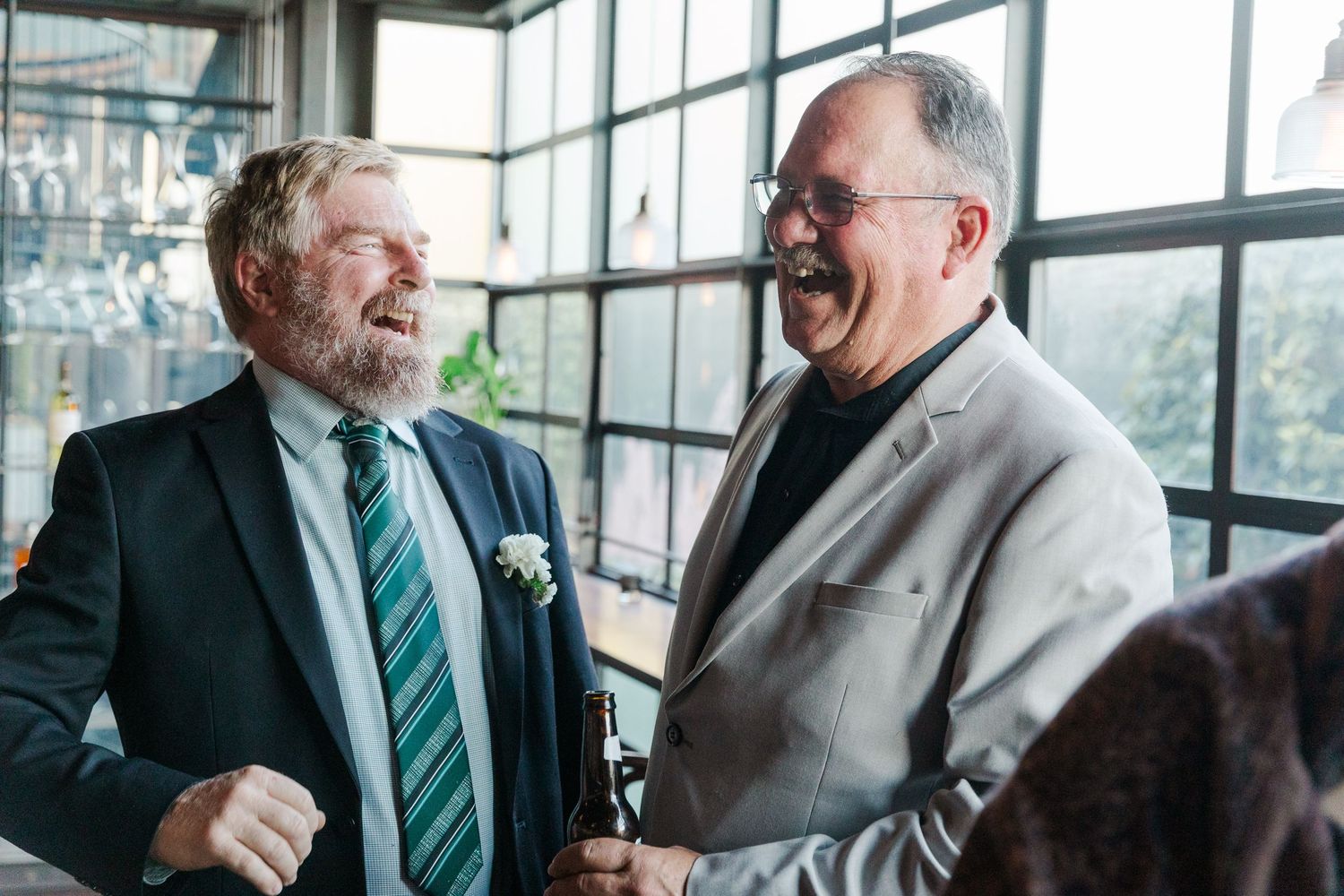 Two older men in suits laughing and conversing joyfully at an indoor social gathering or wedding reception.