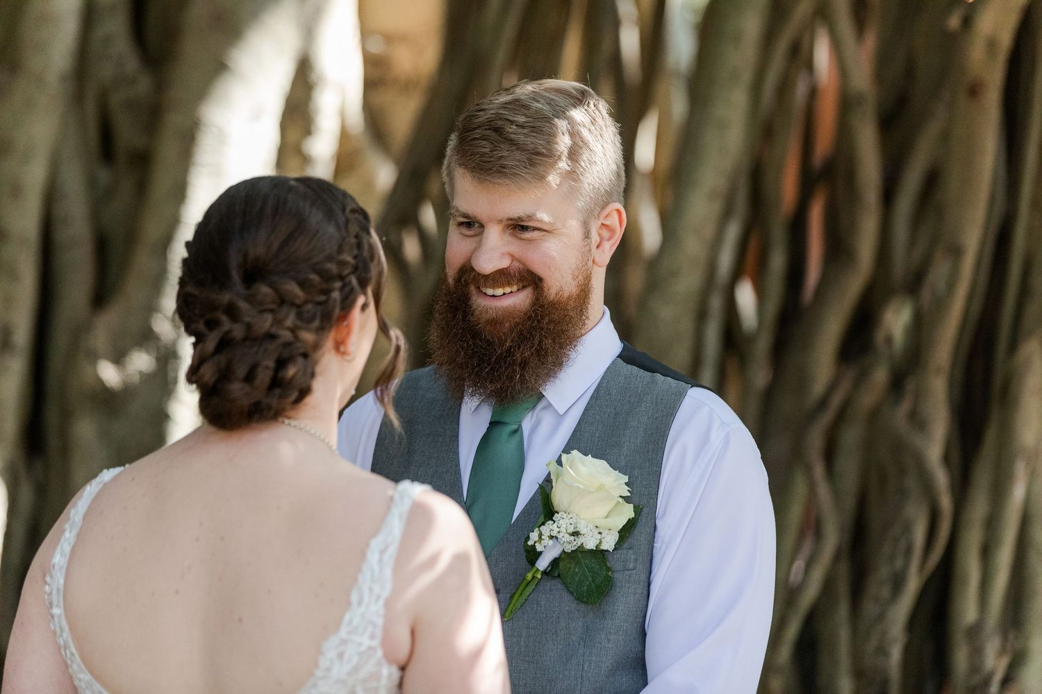 Smiling groom with red beard in gray vest and green tie exchanges vows with bride outdoors at wedding ceremony.