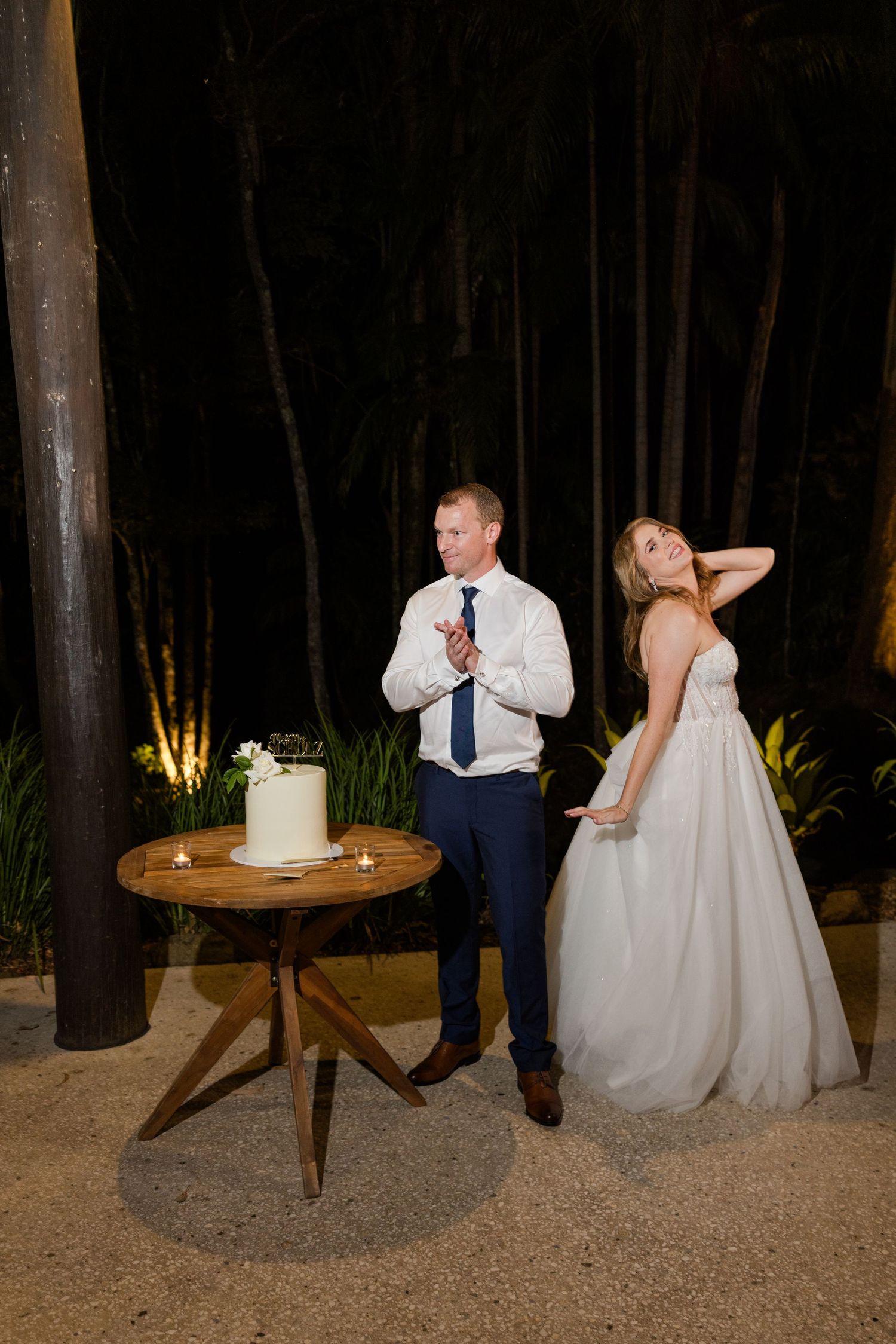 A wedding couple poses playfully next to their white wedding cake on a wooden table in a romantic outdoor forest setting.