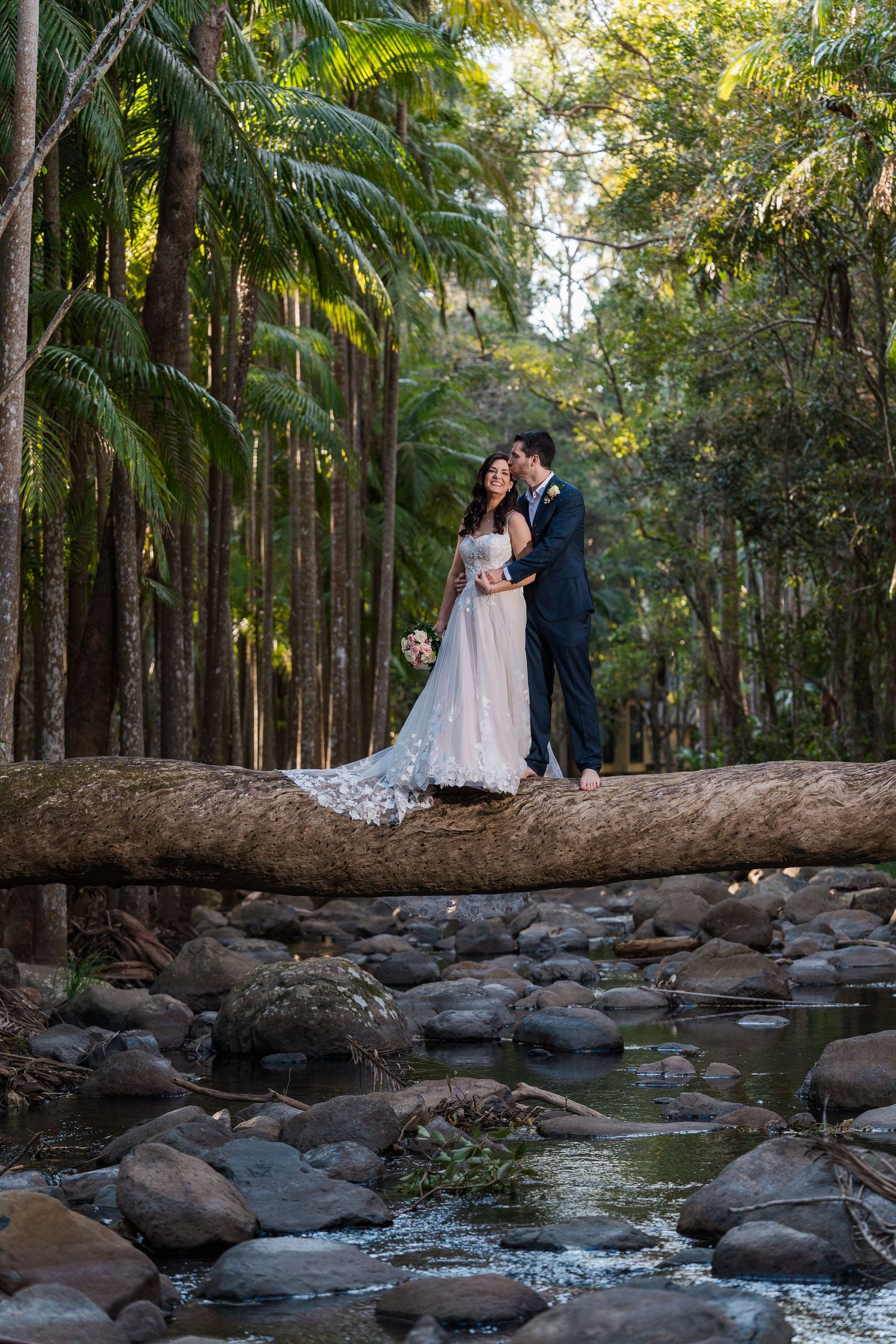 A couple shares an intimate moment on a fallen log over a rocky creek in a lush tropical forest setting.